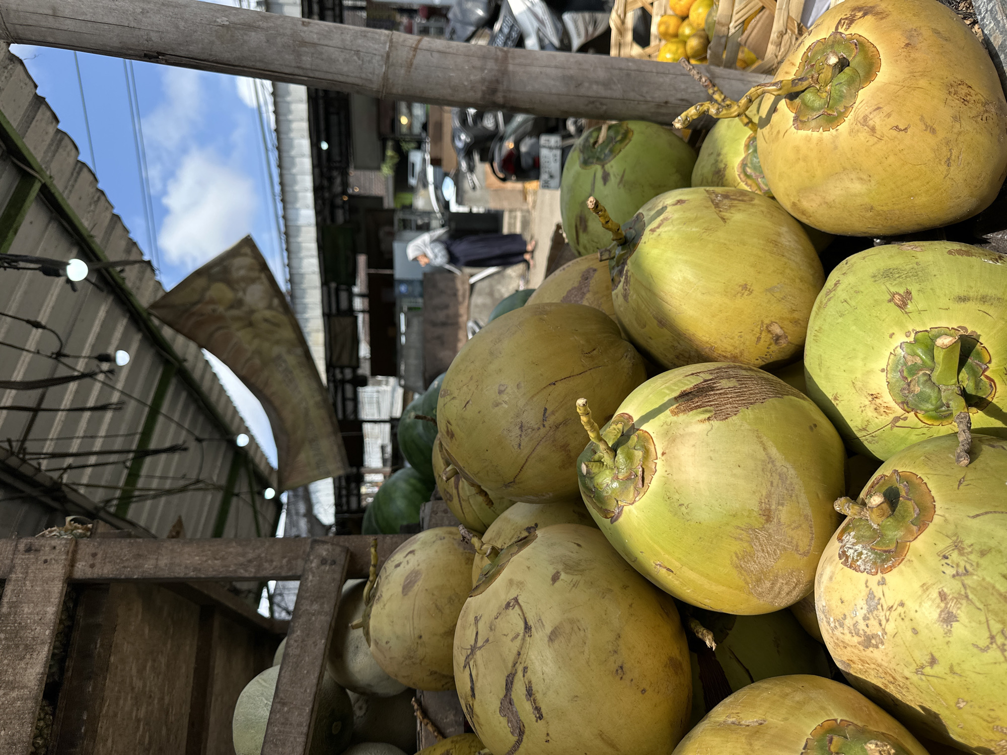 In Kuta Selatan, Indonesia, a roadside stall displays a large pile of fresh coconuts. The coconuts, a mix of green and yellow hues, are stacked in a wooden crate and on the ground. Some coconuts are partially obscured by others, creating a dense visual. In the background, a woman wearing a hijab and dark clothing stands near a scooter, suggesting a bustling market environment. The stall is covered by a corrugated metal roof, with exposed bulbs hanging from the ceiling. A banner, partially unfurled, hangs from the roof. The scene is illuminated by natural sunlight, casting shadows and highlighting the textures of the coconuts and the surrounding structures. In the far right, a basket overflows with bright orange citrus fruits, likely oranges or tangerines, adding a splash of color to the scene. The overall impression is that of a vibrant and lively outdoor market.