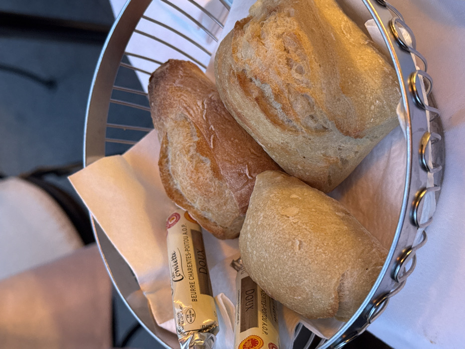 A silver-colored wire bread basket, lined with a white paper napkin, rests on a white tablecloth. Inside the basket are four individual bread rolls, varying in size and crust texture, ranging from light to golden brown. Two individually wrapped portions of "Conviette BEURRE CHARENTES-POITOU A.O.P. DOUX" butter are also visible, one prominently displayed in the upper left, the other partially obscured below the bread. The background is out of focus, showing blurred forms and colors consistent with a dining environment, possibly an outdoor restaurant in an unknown city. No people are clearly visible, and no active interactions are depicted. The scene is static, centered on the food items.
