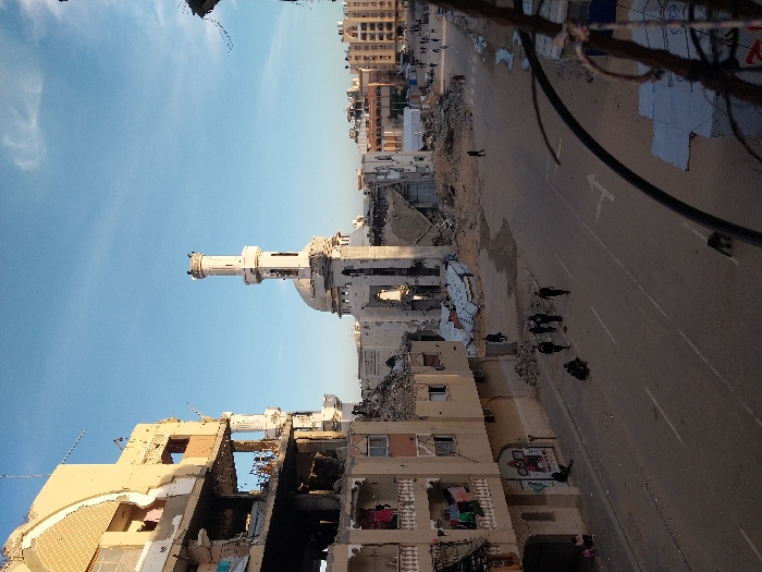The image depicts a war-torn urban environment. Buildings are severely damaged, with visible structural destruction. A mosque's minaret stands relatively intact amidst the rubble. A street is visible, with several figures walking along it. Debris and rubble are scattered across the landscape. The sky is blue, providing a stark contrast to the devastation below. Electrical lines run across the top of the image.