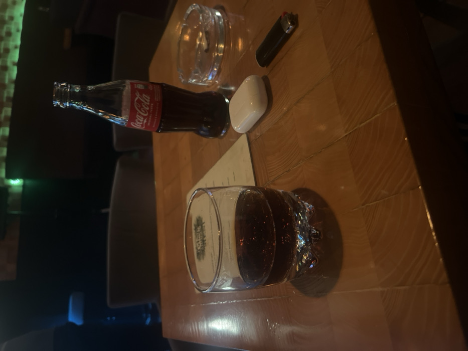 A table with a partially filled glass of cola sits in the foreground. Next to it, a menu card is visible. Further up, a Coca-Cola bottle, a white earbud case, an ashtray, and a lighter complete the collection of items. The table appears to be made of wood, and the image is taken in a dimly lit setting. Several chairs are also visible in the blurred background.