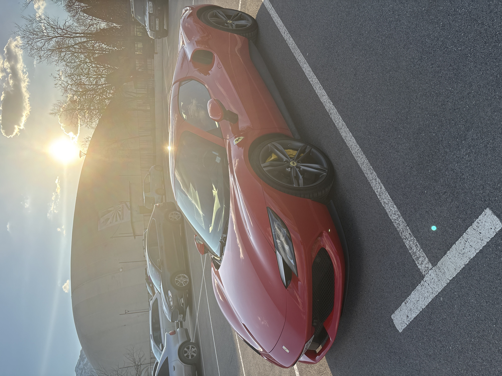 The image shows a red Ferrari parked in a parking lot. The sun is shining brightly, casting a glare on the car's surface. In the background, there are other cars parked and a building. The location is likely in Sofia, Bulgaria, given the presence of what looks like a Bulgarian flag on the building in the background. The car is parked within painted white lines, indicating a designated parking space. The area appears to be an open space, possibly a car park near a building. The car appears to be a later model Ferrari, possibly a 296 GTB or similar. The angle of the photo suggests that it was taken standing.