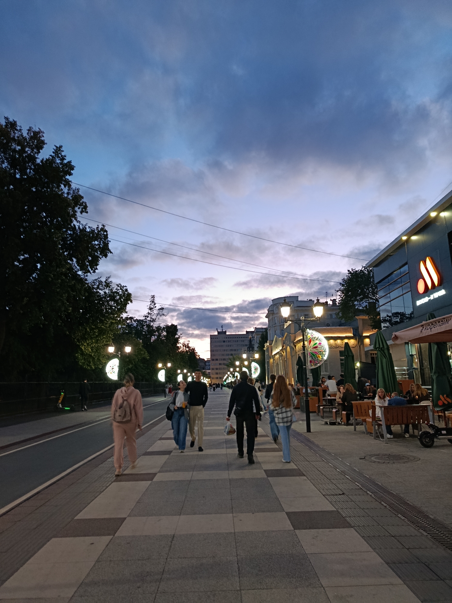 The image depicts a pedestrian street scene in Saratov, Russia, at dusk. The sky is a deep blue with wispy clouds, transitioning to a lighter shade near the horizon. Along the left side of the street, there are large trees and lampposts with circular illuminated decorations. A paved walkway runs alongside the street, bordered by a road with a single lane.
Several people are walking along the pedestrian path. A person wearing a pink tracksuit and backpack is walking away from the camera on the left side of the path. Further down, a couple is walking towards the camera, with the woman holding a drink and the man looking at her. More people are visible in the distance, walking and sitting at outdoor tables.
On the right side of the street, there is a building with a modern facade and large windows. A restaurant or cafe with outdoor seating is visible in front of the building, with green umbrellas providing shade. The tables are occupied by people. The overall atmosphere is that of a lively urban evening.