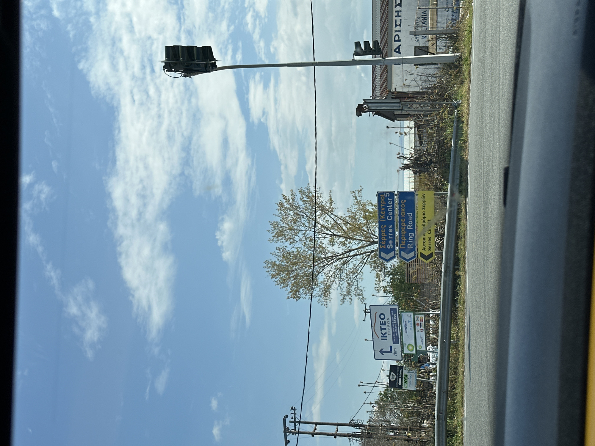 The image is taken from inside a moving vehicle, showing a roadside scene in Lefkonas, Greece. The sky is light blue with scattered white clouds. In the foreground, there is a gray metal guardrail and the edge of the car's dashboard. Beyond the guardrail, there are road signs. One sign indicates "IKTEO" (vehicle inspection center) 7km ahead, along with directions to "Serres Center" and the "Ring Road." Another sign points towards a gas station. Traffic lights are visible overhead. In the background, there are utility poles, trees, and partially visible buildings. The overall environment suggests a highway or main road on the outskirts of town.