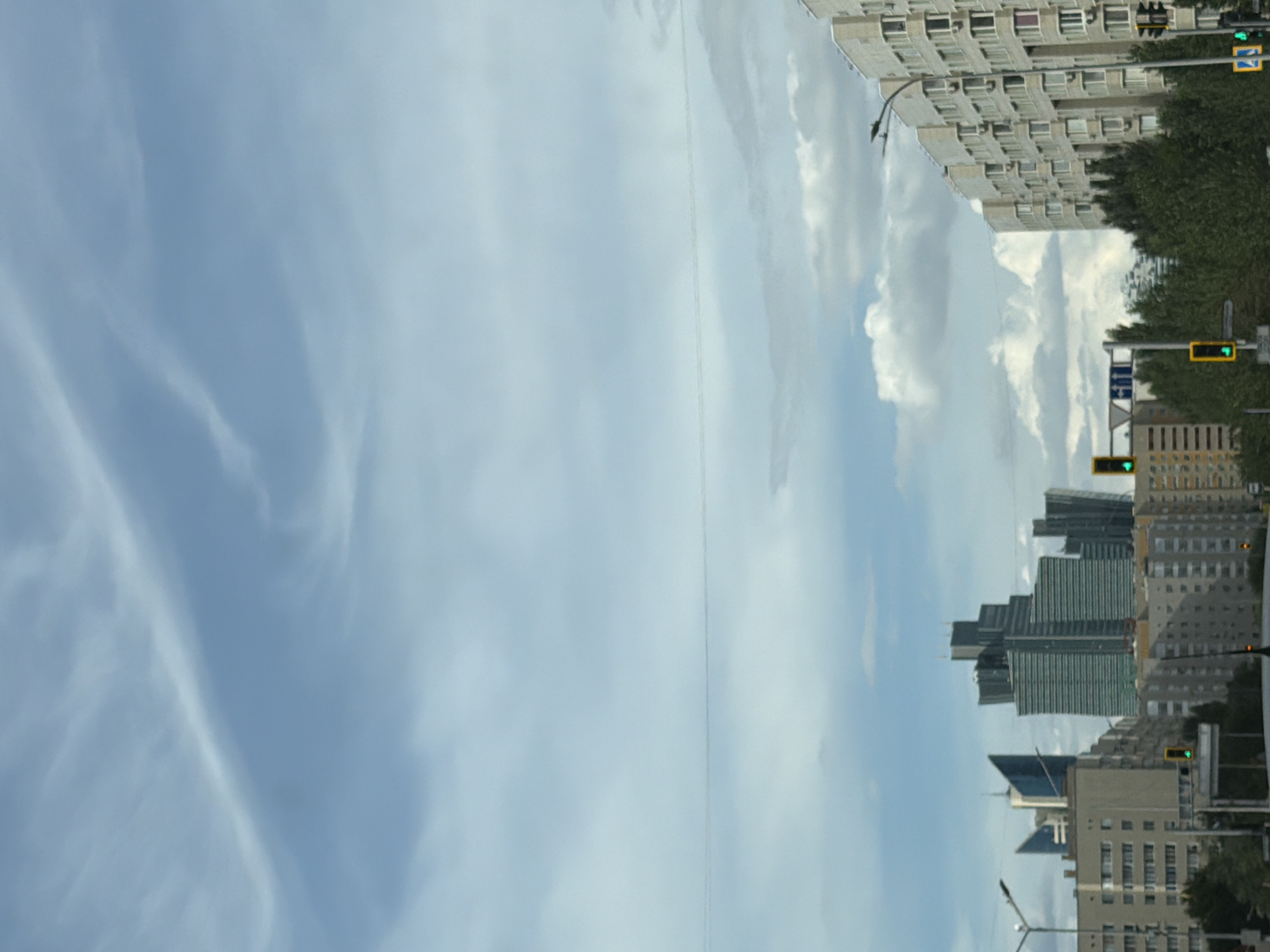 The image depicts a view of the city of Astana, Kazakhstan, with modern skyscrapers dominating the skyline. In the foreground, there are several multi-story apartment buildings, a traffic light with a green signal, and trees. The sky is partly cloudy with a light blue hue. The architectural styles suggest a contemporary urban environment.