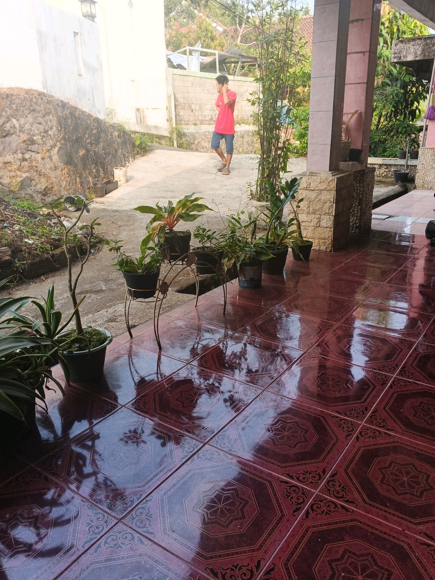 A young man in a red shirt and blue shorts stands on a concrete pathway in front of a tiled patio, talking on his phone. The pathway is bordered by a low stone wall on the left and a residential building with a white facade on the right. Several potted plants are arranged on the patio, which has a glossy, maroon-colored tile floor with an octagonal pattern. A bamboo plant grows near the building's exterior. The scene appears to be a residential area in Keji, Indonesia, likely during daylight hours given the bright illumination and shadows.