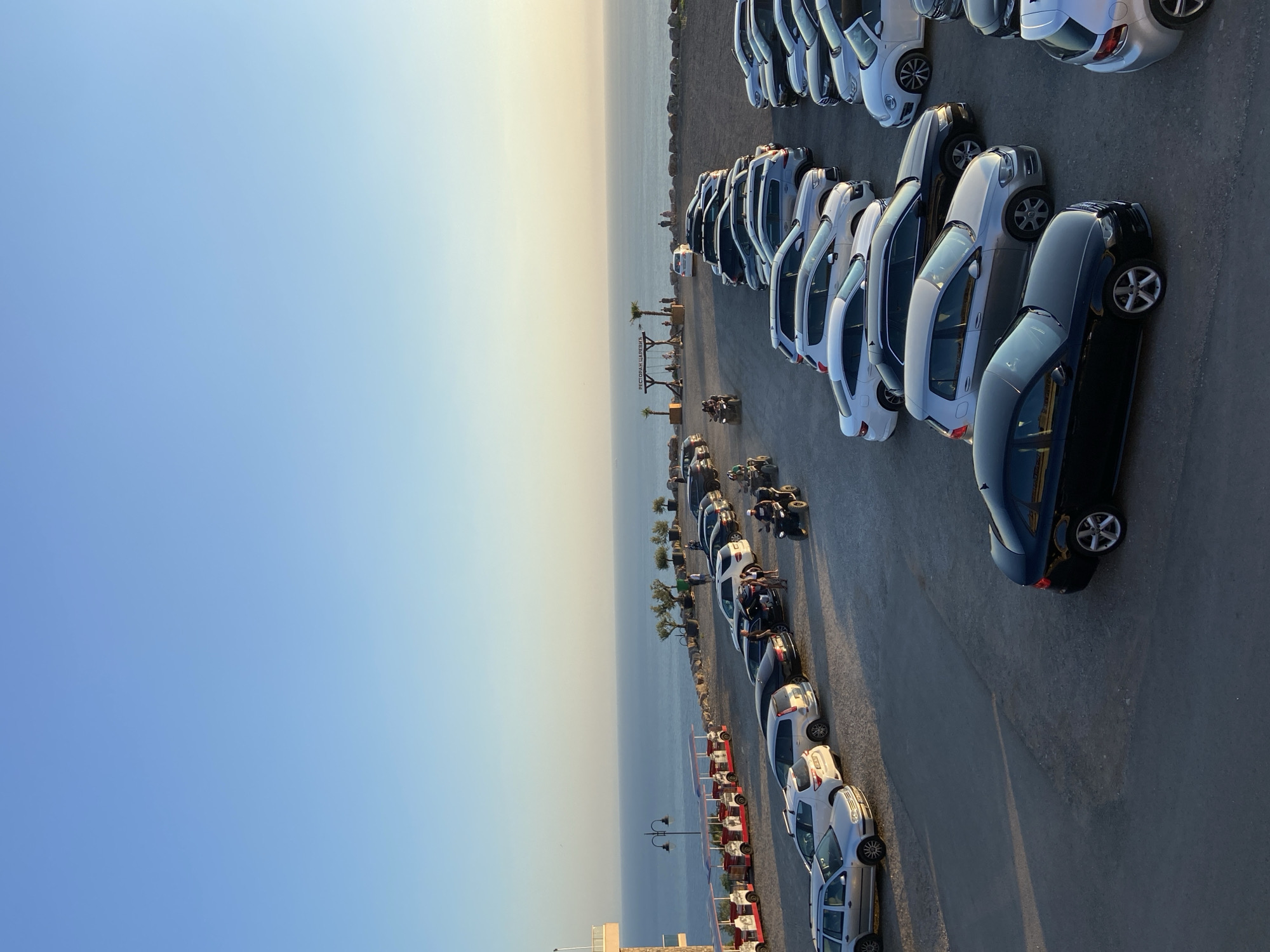 An aerial, high-angle shot captures a parking lot filled with vehicles and a vast expanse of the Adriatic Sea under a clear, pale blue sky. The parking lot is situated on a slight incline, overlooking the sea. Numerous cars are parked in rows, including sedans, SUVs, and a white convertible. Several ATVs are also visible, with two individuals riding one in the middle of the lot. A small, red and white tourist train with multiple cars is parked to the left of the frame. In the distance, a wooden sign with Cyrillic lettering hangs between two palm trees, with a few people gathered nearby. The parking lot's surface is gravelly and uneven. The sea stretches to the horizon, meeting the sky in a faint, hazy line. The overall scene suggests a popular tourist or recreational spot near Kovačko Polje, Montenegro.