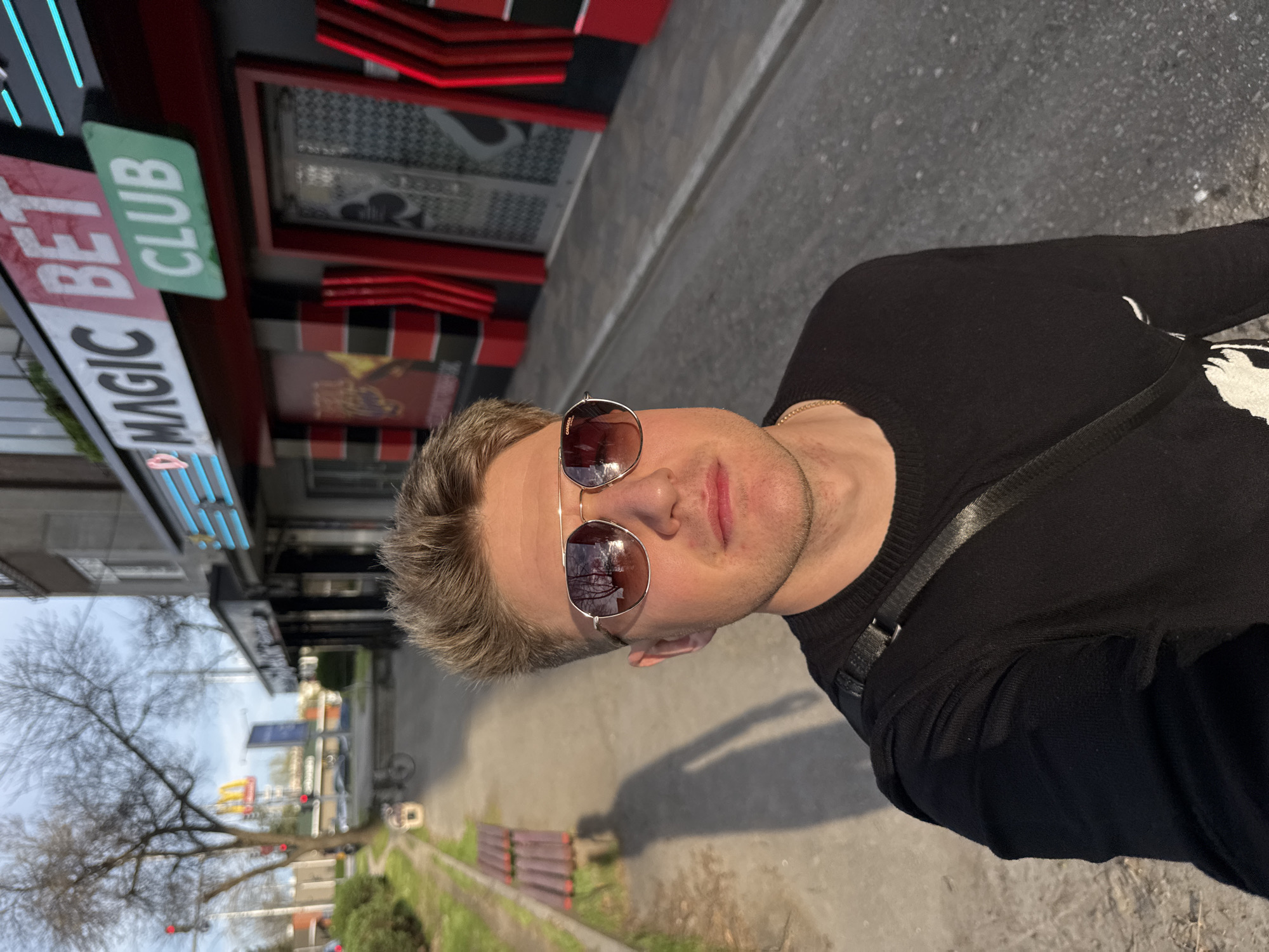 A man wearing sunglasses and a black sweater stands on a sidewalk next to a building bearing the sign "MAGIC BET CLUB." Behind him, the streetscape features a sidewalk, some green vegetation, benches, and in the distance, other buildings. The picture appears to be taken in Plovdiv, Bulgaria.