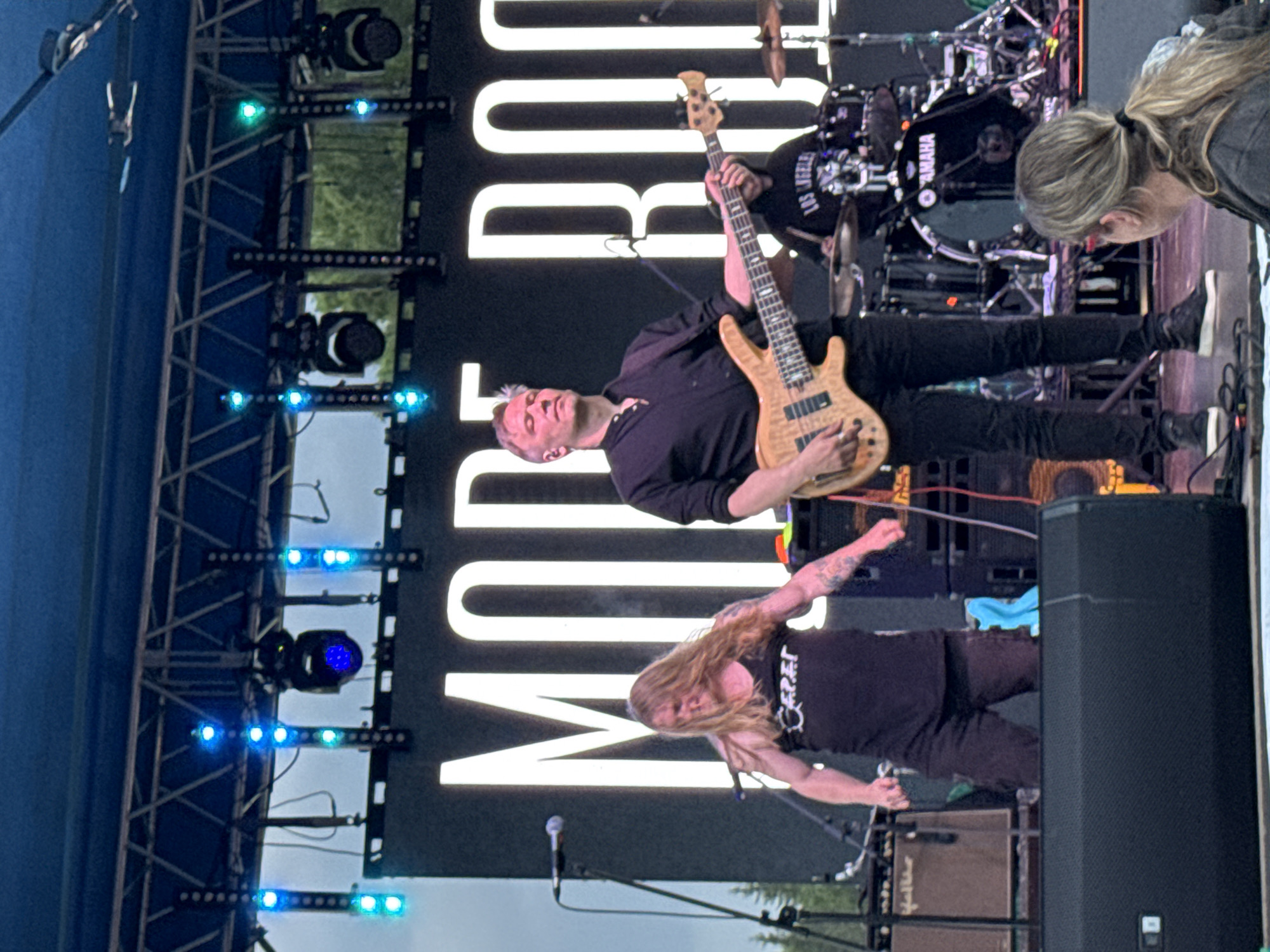 This image captures an outdoor music performance on an elevated stage in Bol'shaya Kivara, Russia.
The scene features a band performing in front of a large black LED screen displaying partial white capitalized text, "MOD ROC," suggesting "MODERN ROCK" or a similar phrase. Overhead, a metal truss structure supports multiple stage lights, including several vertical strips of blue and green LED lights and two dark moving head fixtures, one of which emits a blue light. The sky above is overcast, and trees are visible in the background behind the stage structure.
Three male musicians are visible on stage. On the left, a man with long, light brown hair wears a black sleeveless tank top with a stylized white logo, possibly reading "Ceper." He has visible arm tattoos, is slightly hunched forward, and appears to be moving energetically, with his arms slightly flexed. A microphone on a stand is positioned near him.
In the center, a man with short, light brown and graying hair, wearing a black collared shirt and black pants, plays a light-colored wooden 5-string bass guitar. He is focused on his instrument, with his eyes closed or looking upward. He wears black sneakers with white soles.
Partially visible on the right, behind the bassist, a drummer wearing a black hoodie with "LOS ANGELES" text plays a black Yamaha drum kit, identifiable by the logo on the bass drum. Various cymbals and drum hardware are present. Stage monitors and amplifiers are visible around the performers, including a brown-colored amplifier on the left and a black speaker on the right behind the drummer. A blue cloth or towel is on the stage floor near the center-right.
In the immediate foreground, with her back to the viewer, a woman with long blonde hair tied in a ponytail is seated or kneeling, wearing a dark jacket, observing the performance.
