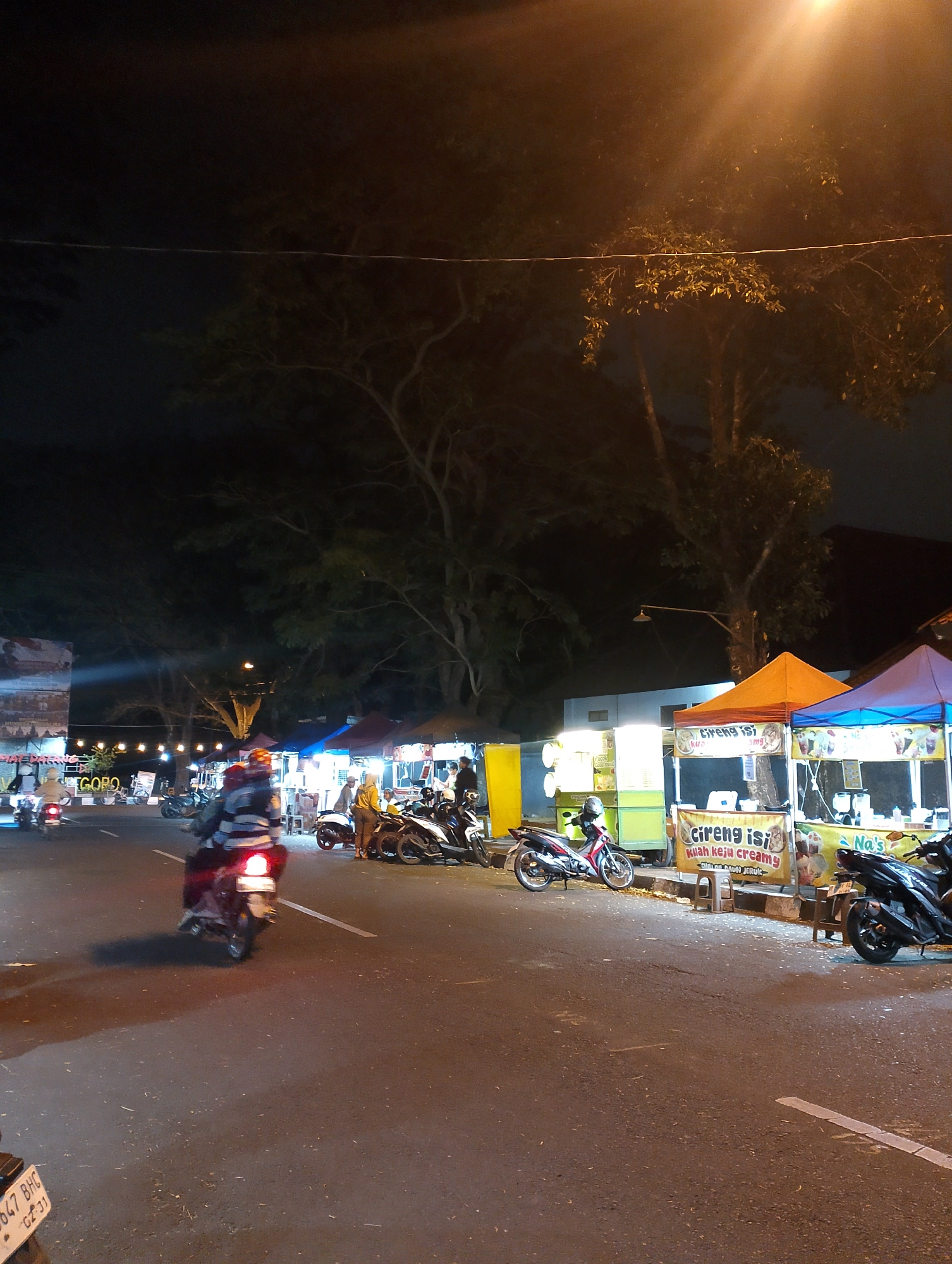 The image depicts a street scene at night in Magelang, Indonesia, likely a bustling area with food stalls and street vendors. The lighting is dominated by artificial sources, including streetlights and illuminated signs from the stalls, casting a warm glow on the scene. Several motorbikes are present, indicating local transportation and the common use of two-wheeled vehicles. Some motorbikes are parked, while others are in motion, with one rider prominently featured in the foreground, their taillight illuminated.
The street itself is paved and appears to be a two-lane road. On the right side of the street, a row of temporary food stalls with colorful awnings are set up. These stalls have signs advertising their offerings, such as "Cireng isi" and "kuah keju creamy," suggesting they sell local snacks and food items. People can be seen standing and interacting around these stalls, likely customers or vendors.
On the left side of the street, more stalls are visible, some with bright lights illuminating their products. A large banner with Indonesian text and images is also present on the left, potentially advertising an event or a business. Trees with dense foliage line the background, creating a natural backdrop against the urban setting. The overall atmosphere appears lively and characteristic of a night market or a commercial street in an Indonesian city.