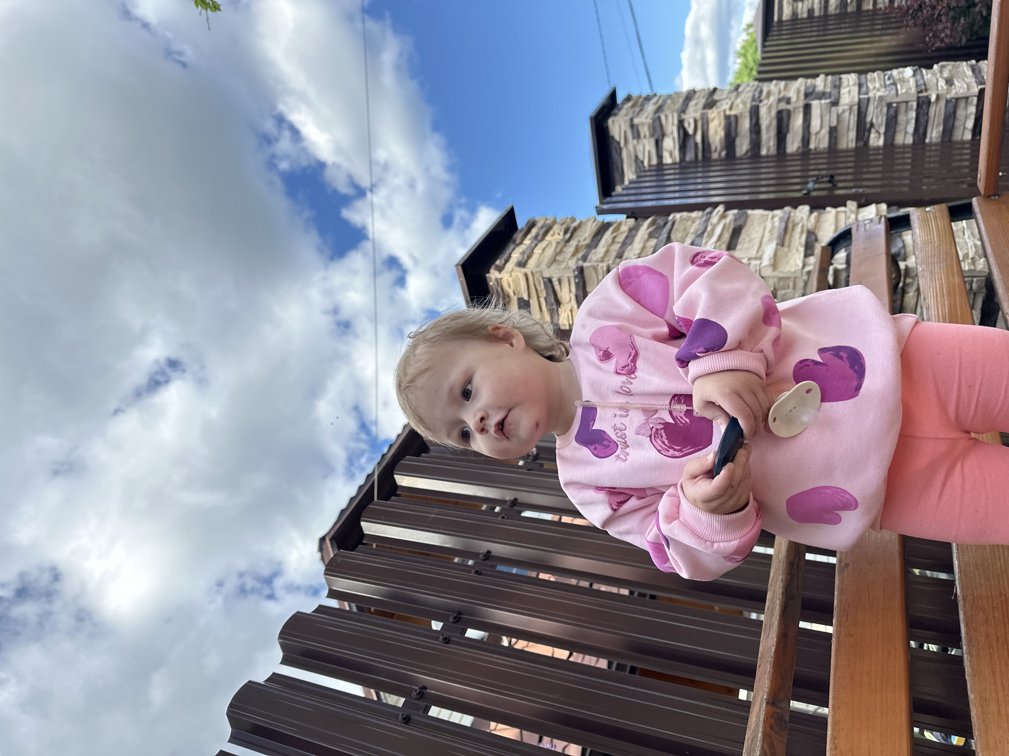 A young child, likely a toddler, is captured from a low-angle perspective in an outdoor setting. The child wears a light pink long-sleeved top featuring large purple heart-like designs and matching pink pants. On the chest of the top, the text "Trust in Love" is visible alongside a heart motif. The child has blonde hair and a calm, slightly curious expression, looking slightly upwards and to the viewer's left. They are holding a small black object in one hand and a white pacifier in the other.
The background features a bright blue sky dotted with scattered white clouds, indicating a clear, sunny day. Behind the child are structures with textured, layered walls, possibly imitation stone or brick, topped with dark brown slatted sections that resemble a fence or railing. A wooden structure, potentially part of a play area or outdoor seating, is visible in the foreground, where the child appears to be positioned. The scene suggests a relaxed moment of childhood observation or play in an outdoor location, possibly a backyard or a public area in Nizhny Novgorod, Russia.