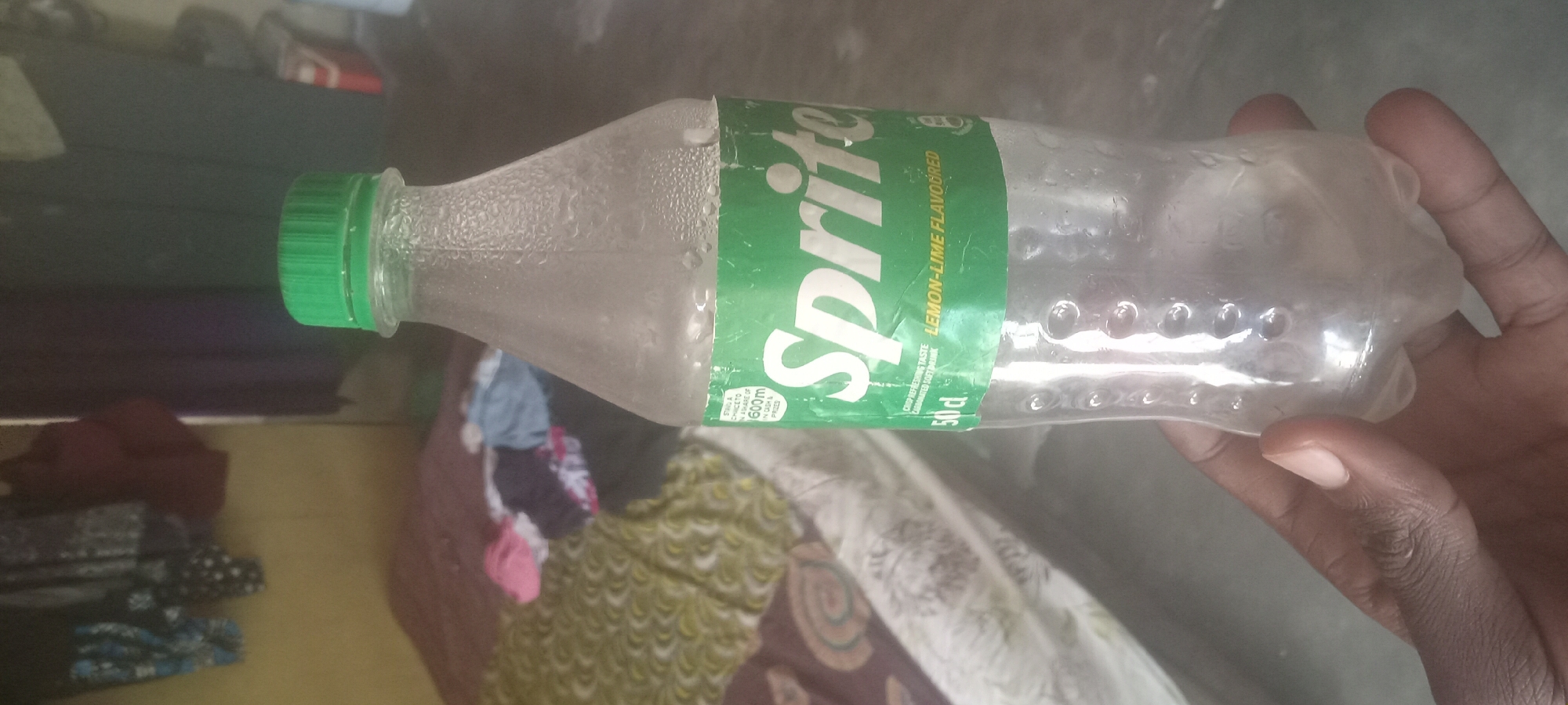 An empty, partly-condensated, 50cl plastic Sprite bottle with a green cap is held horizontally in a person's hand. The bottle label indicates "Lemon-Lime Flavoured". The bottle is resting at an angle on a surface that appears to be a bed or floor, amidst some patterned fabric or clothing. The image was likely taken indoors. No indication of the city of Mbribit, Nigeria, is present in the image.