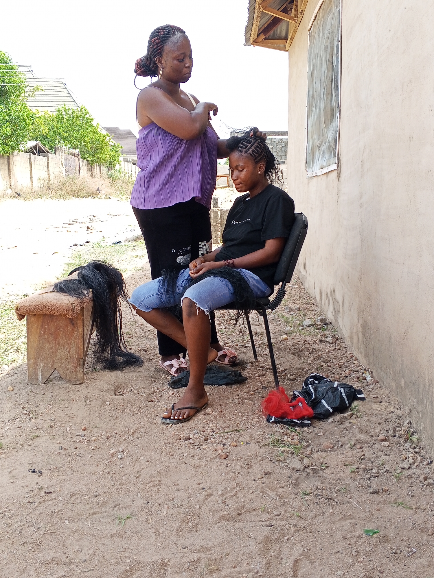 This outdoor photograph, taken in Adonkolo, Nigeria, depicts two Black women engaged in hair braiding under bright daylight. A woman in a black t-shirt and ripped blue jeans is seated on a black office-style chair, her head partially braided with cornrows, while long black hair extensions rest on her lap. Another woman stands behind her, wearing a purple pleated top and dark pants, meticulously braiding the seated woman's hair. Her own hair is styled in braids.
To the left of the frame, a wooden stool with a brown, padded top holds additional black hair extensions. The ground is unpaved, consisting of dirt and small gravel. In the background, a light-colored building with a window covered by a mesh screen is visible, along with a fence and green foliage, suggesting a residential or communal outdoor area. A black plastic bag with white stripes and some red material lies on the ground near the seated woman's feet. Partial text "C O NT" is visible on the standing woman's pants. Both individuals appear focused and calm, indicating the typical, time-consuming nature of hair braiding. The bright lighting suggests it is daytime with clear weather.