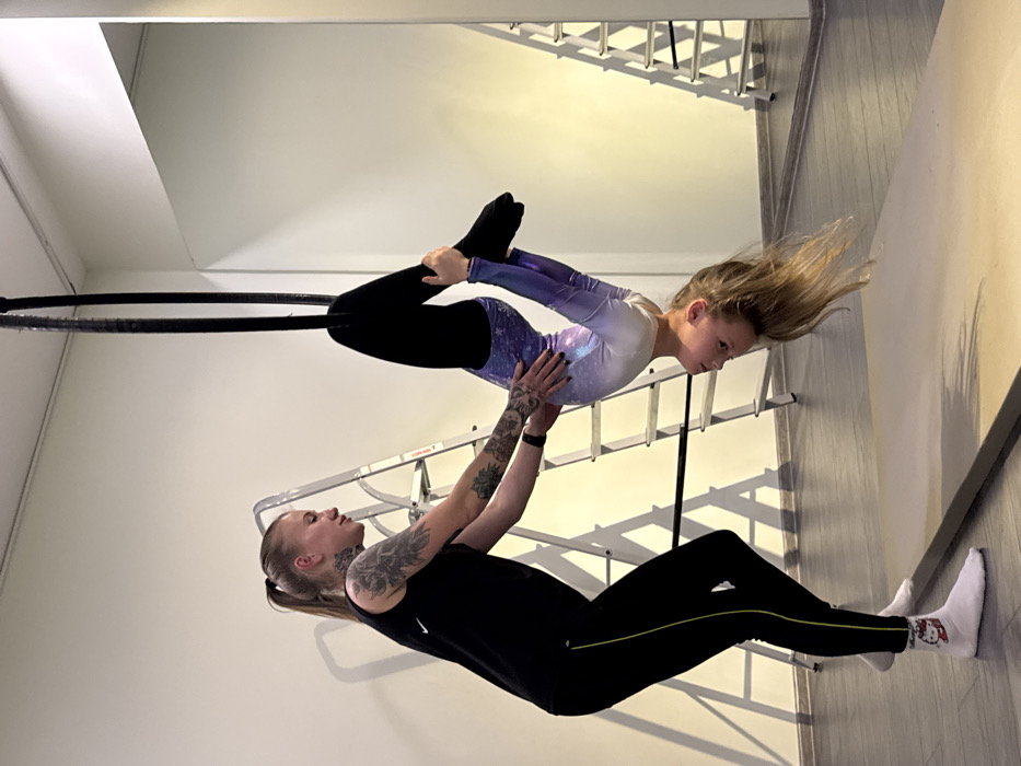 An aerial acrobat is shown holding a girl performing a maneuver on an aerial hoop. The girl wears a celestial leotard, black leggings, and black socks, while her long blonde hair streams behind her. The tattooed acrobat wears a black Nike top, black leggings with a yellow stripe, and white socks. The image is captured in a studio with a mirror and a ladder visible in the background.
