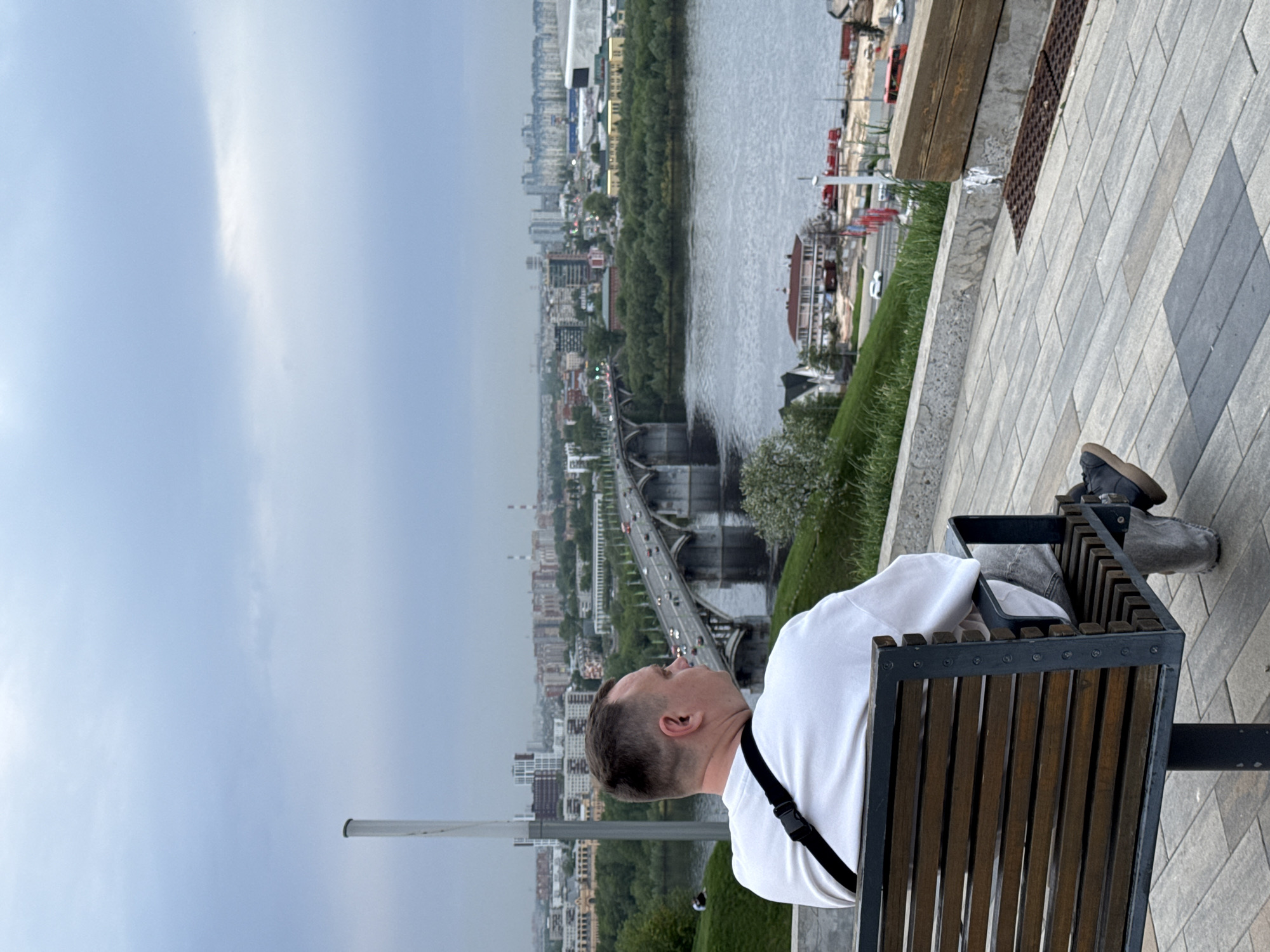 A man sits on a bench overlooking a river and bridge in Nizhny Novgorod, Russia. He is wearing a white shirt and has a black strap across his body. The bridge spans the river, connecting parts of the city. The cityscape includes buildings and greenery, with an overcast sky. The bench is made of wood and metal. The area around the bench is paved with patterned stones.
