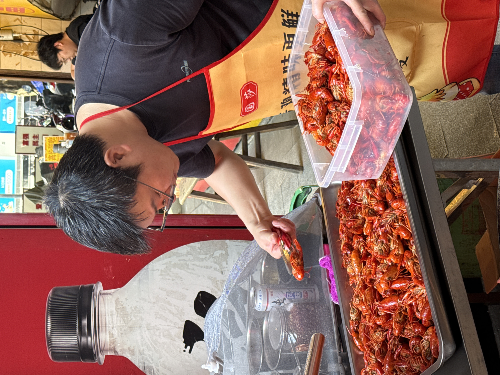 A person wearing glasses, a black t-shirt, and a tan apron with red trim is pouring cooked crawfish from a clear plastic container into a metal tray overflowing with crawfish. The apron has Chinese characters on it. The person is standing next to a counter with a red background featuring a bottle design. Behind the counter is an area containing cooking utensils and cans. A second person is partially visible in the background wearing glasses and a dark shirt. Based on the markings on the can, the city is Beibei in China.