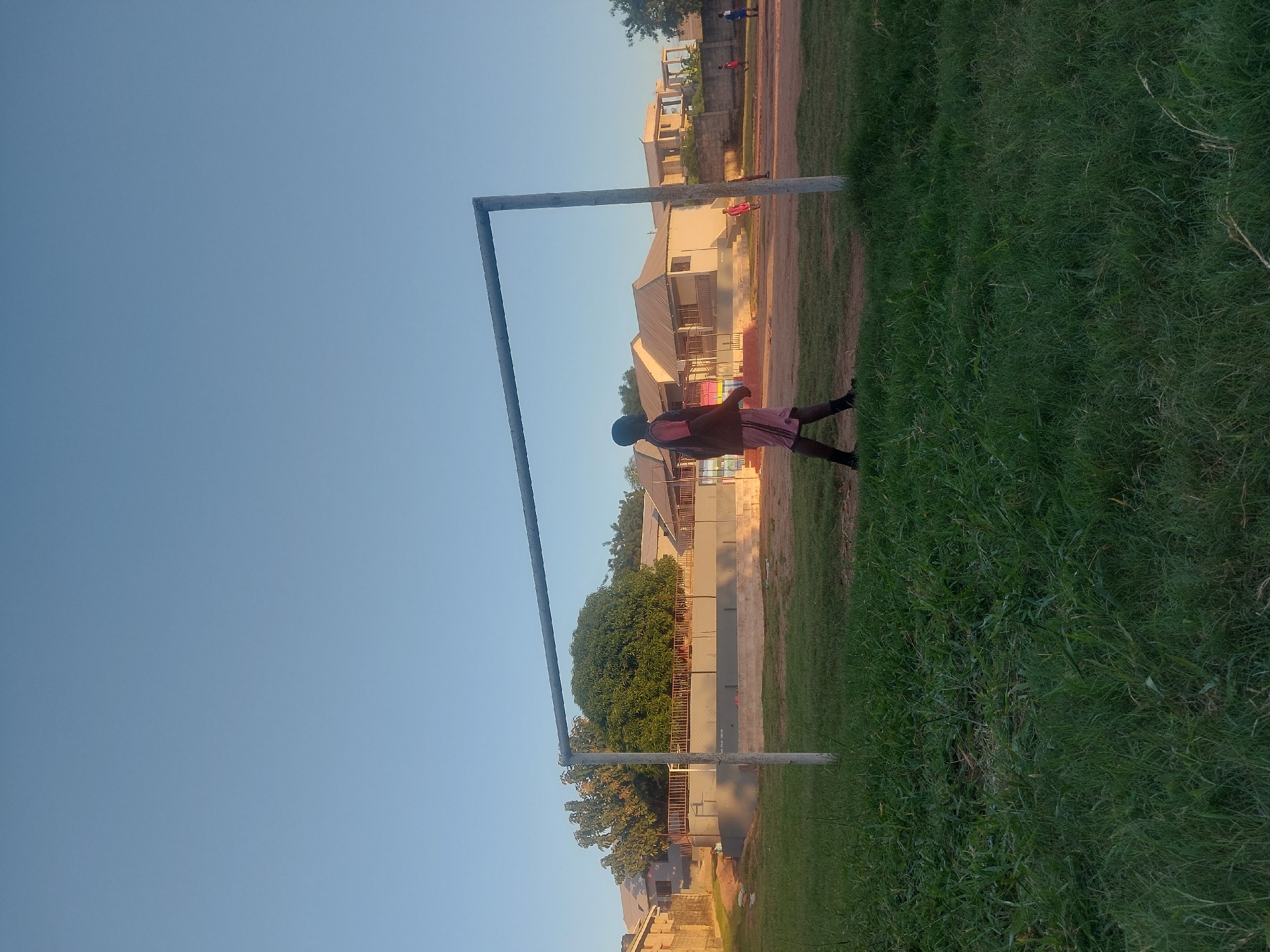 The image depicts a young person standing on a grassy field next to a soccer goalpost. The location is likely an urban area within Dar es Salaam, Tanzania, as evidenced by the presence of houses in the background. The sky is clear and blue. The individual appears to be wearing dark shoes, light pink shorts and a brown shirt, and is facing right in the photo. There is green grass with some brown dirt/mud next to it, leading to a house.