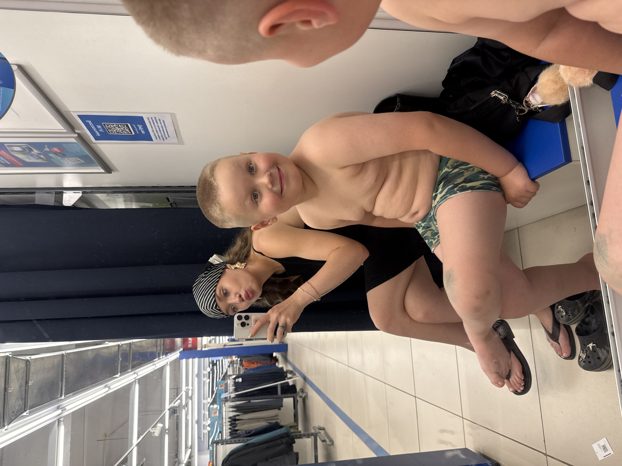 In a brightly lit fitting room, a young boy with blond hair and blue eyes smiles at the camera while wearing camouflage-patterned swim trunks. His skin is slightly red, possibly from sun exposure. Next to him, a woman with blond hair, wearing a black dress and a black and white striped headscarf, takes a selfie with her phone. She has a ring on her left hand and a bracelet on her right wrist. Her expression is playful, with her lips pursed. Both are barefoot, with the woman wearing black flip-flops and the boy wearing black Crocs. Behind them, a black bag and a fuzzy toy are visible. The room appears to be part of a retail store, with racks of clothing visible in the reflection of a large mirror that covers much of the wall. One sign in the mirror has a QR code and text in Russian. The overall mood is casual and lighthearted, suggesting a fun shopping trip or perhaps trying on swimwear. The setting is likely a store in Votkinsk, Russia.
