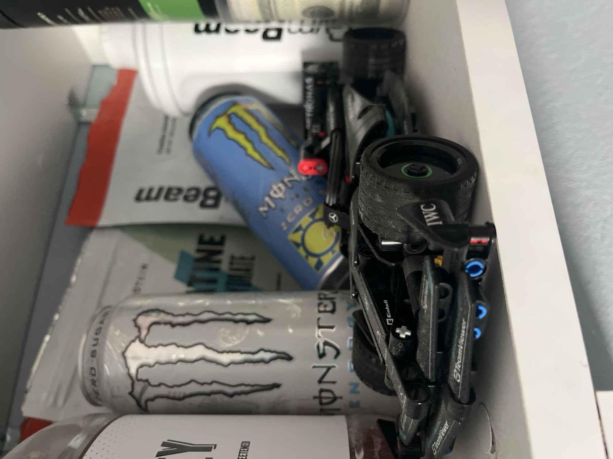 The image shows a drawer filled with various items. There are two cans of Monster energy drink, one silver and one blue, along with a white bottle labeled "beam" and a white container. A toy race car with "IWC" and "TeamViewer" printed on it sits among the items. The surrounding environment is a white drawer. The location is unknown and cannot be determined from the image. No people, subjects, actions, or interactions are depicted. The image does not provide information about the city of Pokrovnik, Bulgaria.