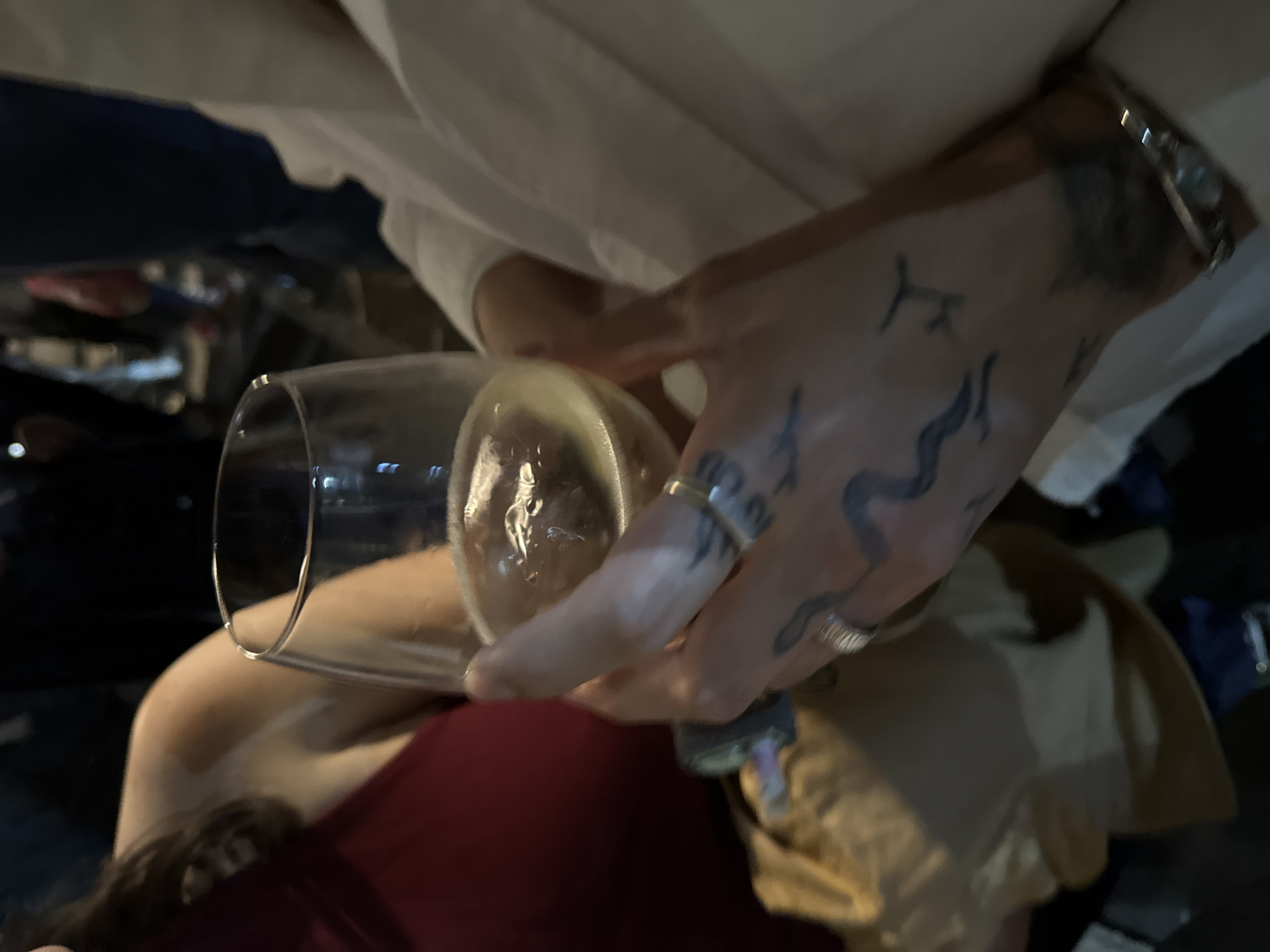 A tattooed hand holds a partially filled wine glass. A second hand steadies the glass. The location is possibly Yerevan, Armenia, but there is no indication in the image to confirm this. A person with a red shirt and hair is in the foreground, while two other people are visible in the background. The background is dark.