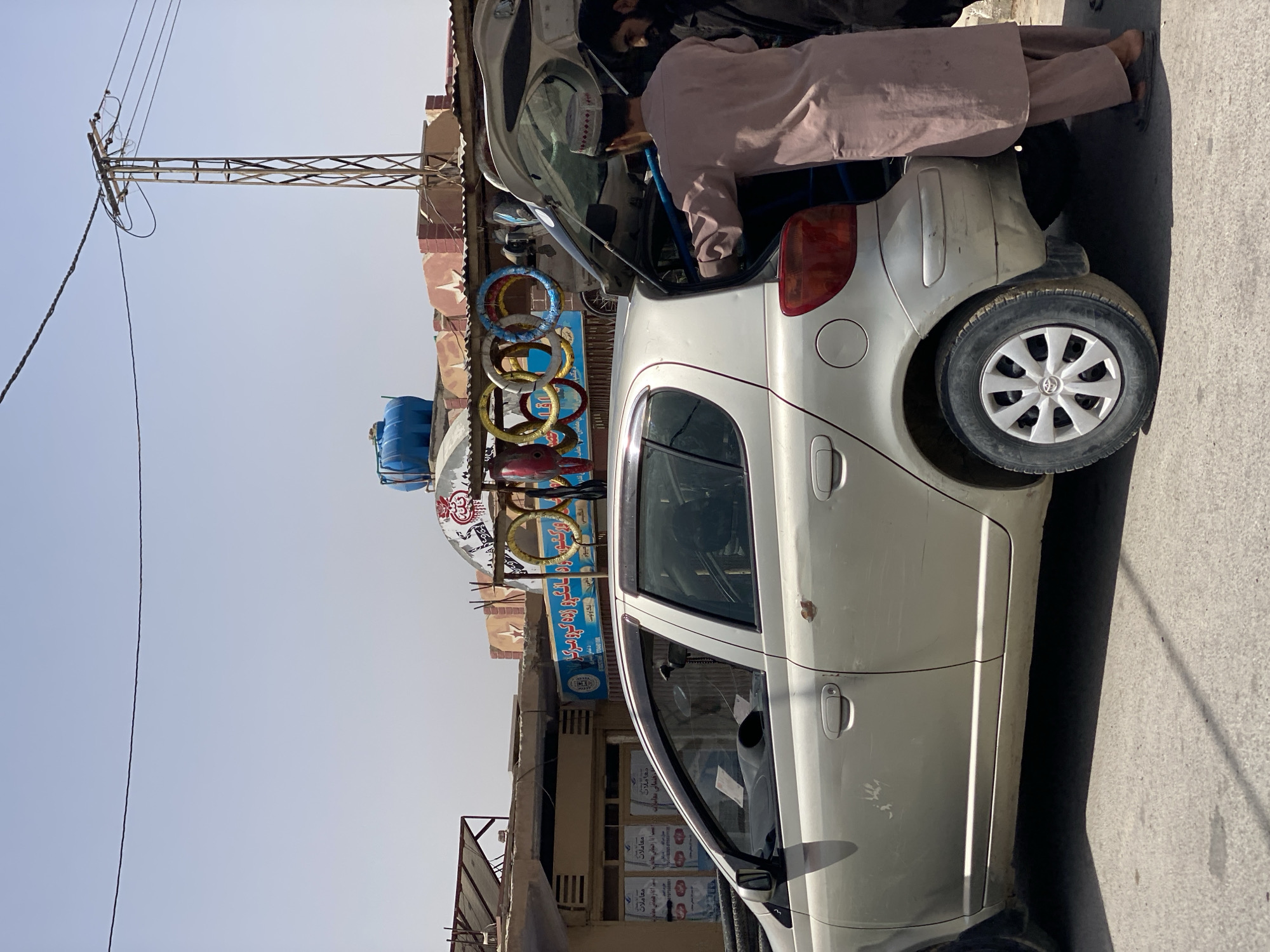In Kandahar, Afghanistan, a silver Toyota sedan is parked on a street, with its trunk open. Two men are present. One man, wearing a light brown shalwar kameez and a white and black patterned cap, is leaning into the open trunk, appearing to place or retrieve an item. The second man, to the right of the first, is wearing dark clothing and looking towards the trunk. Behind the car, a building with a signage featuring text in Dari and English is visible. The building's roof has colorful circular objects mounted on it, resembling Olympic rings. A utility pole with wires stands in the background, and the sky is clear and bright. The car has visible wear and tear, including rust spots and scratches on its exterior. The ground is paved with asphalt.