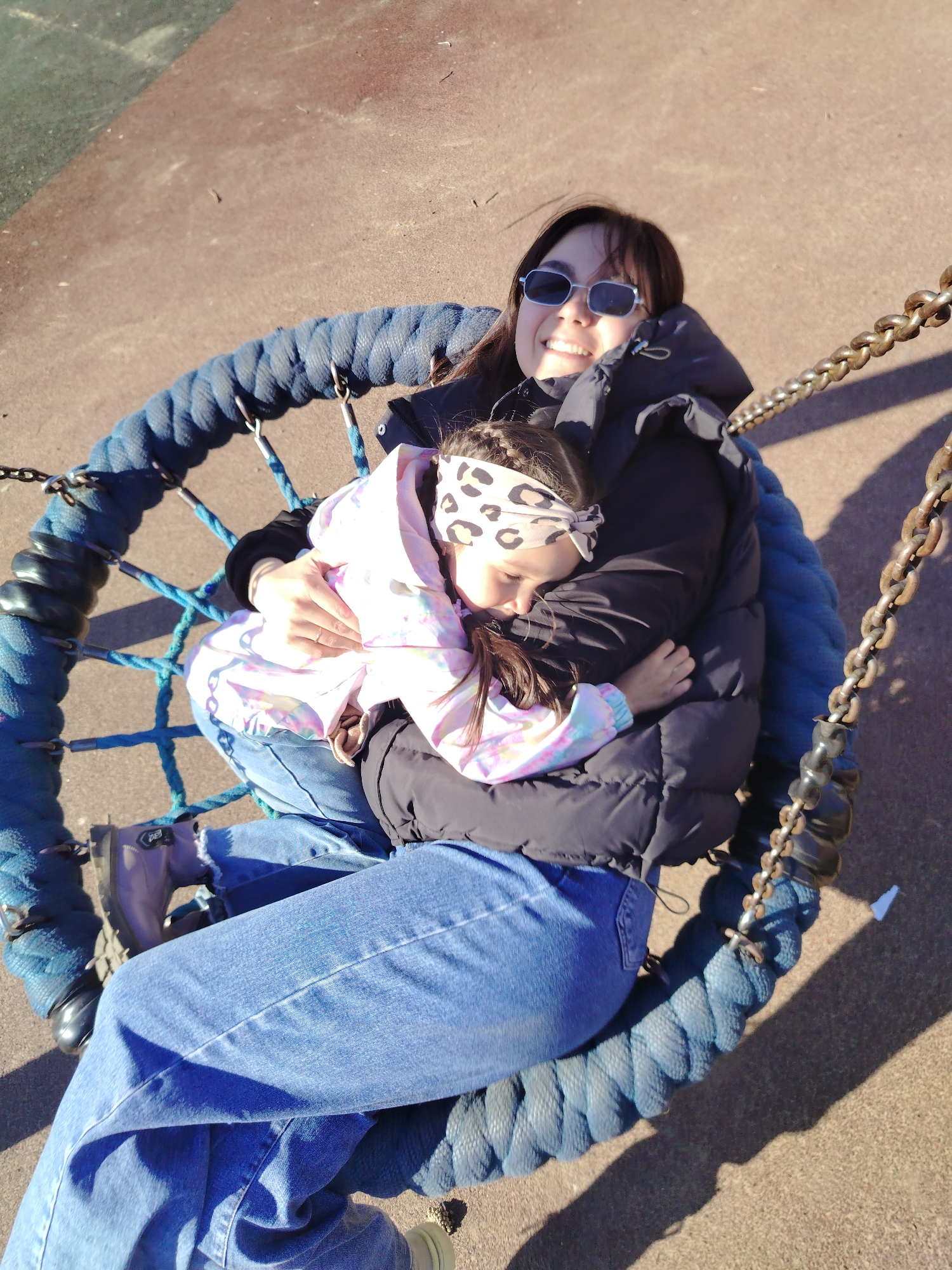 The image captures an adult and a child sitting together in a large, blue, basket-style swing, likely in an outdoor playground or park in Noviy, Russia. The adult, positioned in the foreground, has long dark hair and wears dark sunglasses, a black puffer jacket, and blue jeans. She smiles warmly at the camera. Cradled in her arms is a child with dark hair, wearing a light-colored, possibly tie-dye patterned jacket and a leopard print headband. The child appears to be resting or asleep, with their eyes closed, snug against the adult.
The swing itself is made of thick blue rope, forming a circular nest, and is suspended by heavy metal chains. Bright sunlight illuminates the scene, casting distinct shadows from the chains and individuals onto the reddish-brown, possibly rubberized, ground surface. The overall atmosphere conveys a sense of peace, comfort, and affection between the two individuals. There is no discernible text visible in the image.