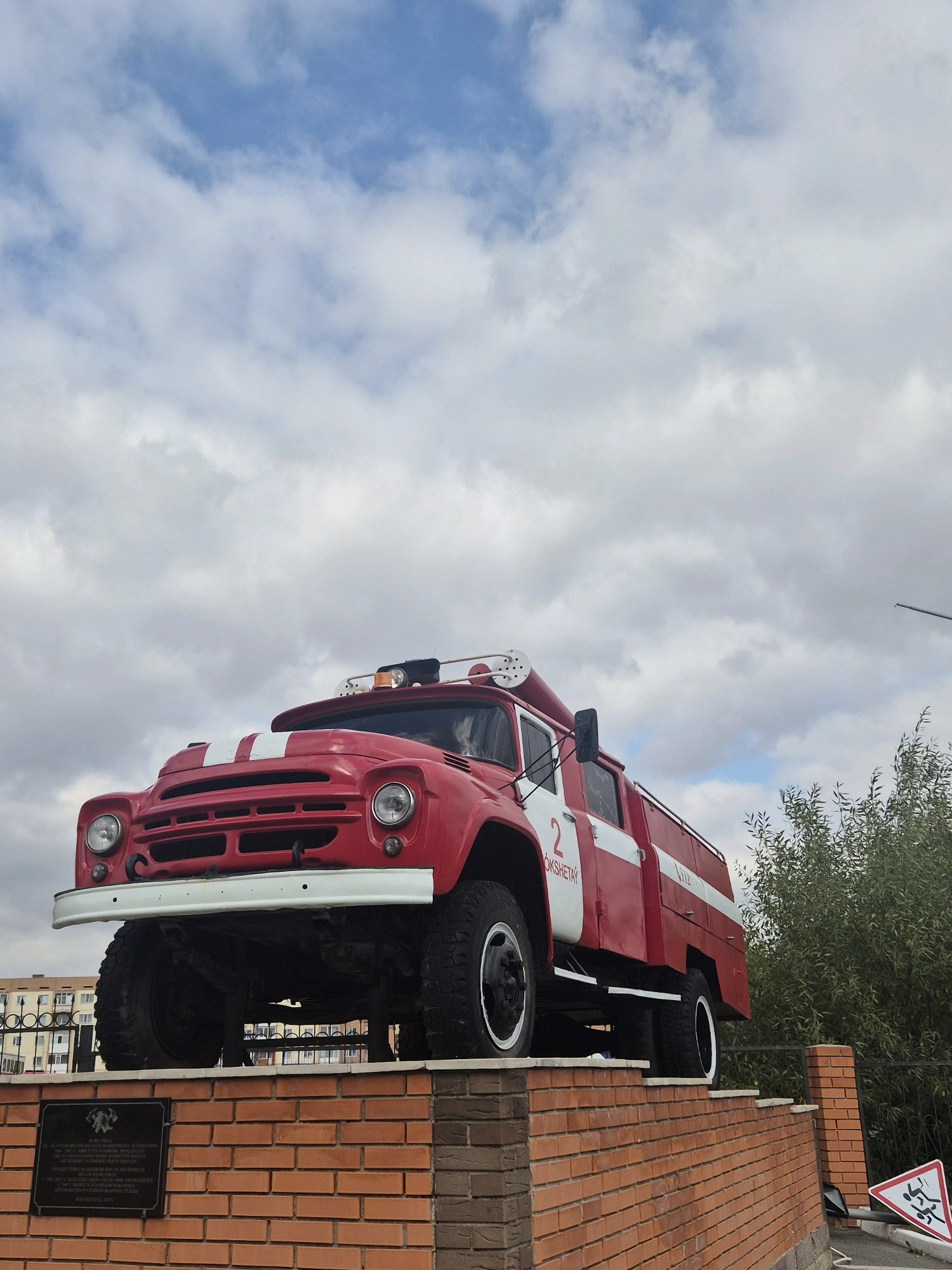 A bright red vintage fire truck, possibly a ZIL-130, is displayed on a brick pedestal outside. The truck has white racing stripes on the hood and white trim along the side. The number "2" and Cyrillic text reading "KOKSHETAY" are visible on the side. The fire truck is positioned at an angle, with its front facing left and its side visible. It is adorned with a light bar and what appears to be a hose reel. The pedestal is made of red brick and has a plaque at its base, detailing information about the fire truck's history. A fence and lush green trees are visible behind the truck, and a distant building can be seen on the left. The sky above is filled with dramatic, fluffy white clouds. The overall scene suggests a monument or memorial dedicated to firefighters in the city of Kokshetau, Kazakhstan.