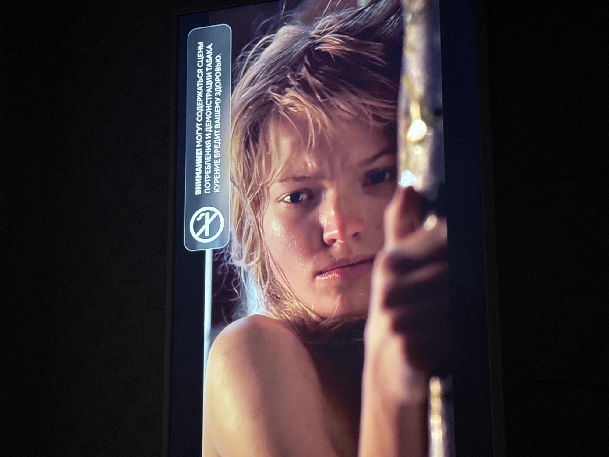 The image shows a close-up of a woman's face, partially obscured by a vertical object. She has fair skin, light-colored, tousled hair that appears wet, and an intense gaze. Part of her upper torso and shoulder are visible, suggesting she may be unclothed or wearing minimal clothing. To the left of the woman is a rectangular warning label in Russian text, containing the symbol of a crossed-out cigarette. The background appears dark and undefined. The image appears to be taken from a television or digital display. There is no indication of the location of Mytishchi, Russia, in this image.