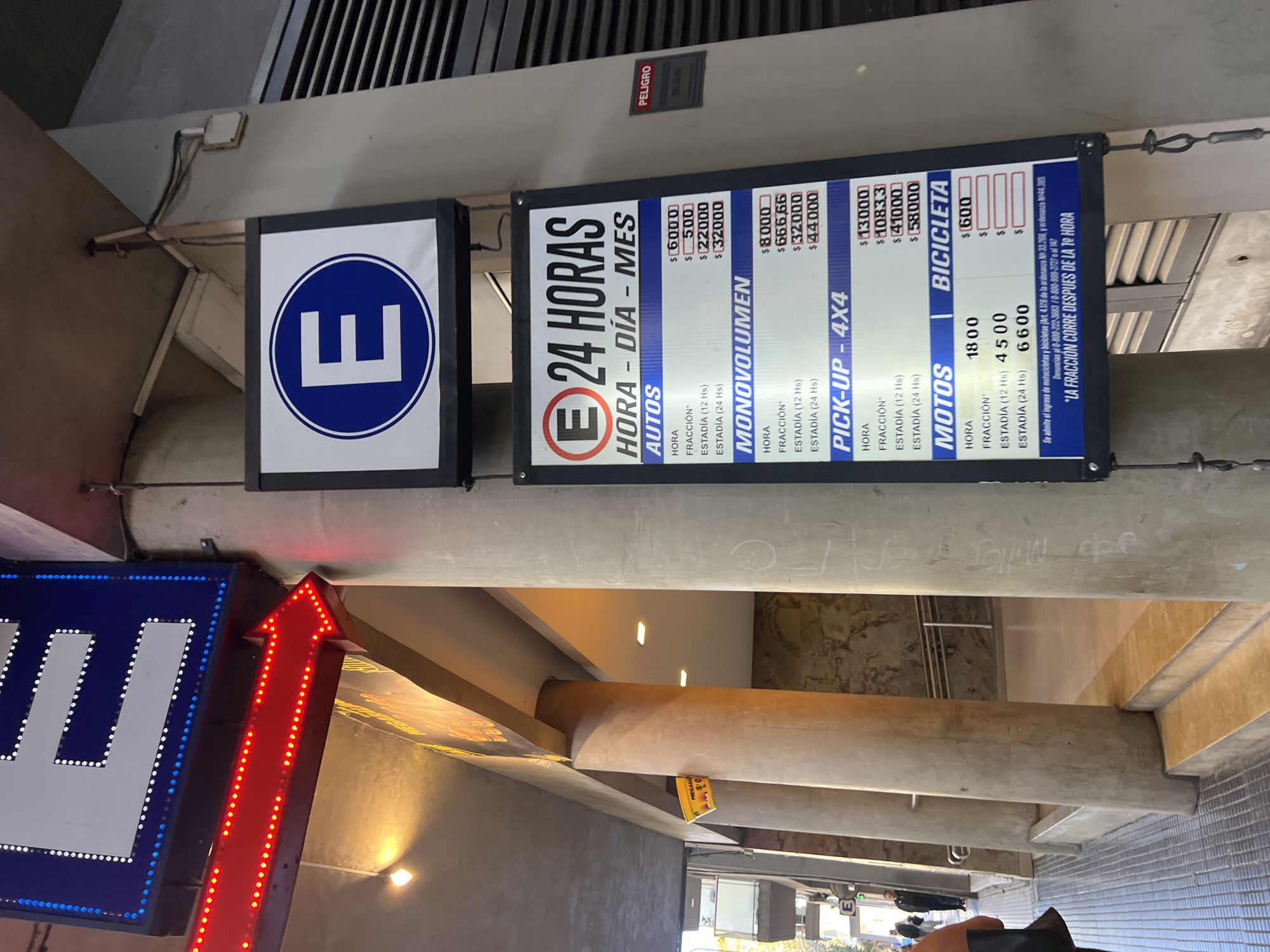 The image captures a detailed view of signage at the entrance or within a parking facility in Buenos Aires, Argentina. The primary subjects are several signs providing information about parking services and rates.
Dominating the left side is a large blue and white circular sign featuring a white "E," commonly indicating "Estacionamiento" (Parking). Adjacent to it, a rectangular digital display sign, also featuring an "E," advertises "24 HORAS" (24 Hours) and "HORA - DÍA - MES" (Hour - Day - Month), indicating continuous operation and various payment options. This sign provides a comprehensive price list for different vehicle types:
- **AUTOS (Cars):** $6000 (Hour), $500 (Fraction), $22000 (12 Hs Stay), $32000 (24 Hs Stay).
- **MONOVOLUMEN (Minivan/SUV):** $8000 (Hour), $666.66 (Fraction), $32000 (12 Hs Stay), $44000 (24 Hs Stay).
- **PICK-UP - 4X4 (Pickup - 4x4):** $13000 (Hour), $1083.3 (Fraction), $41000 (12 Hs Stay), $58000 (24 Hs Stay).
- **MOTOS (Motorcycles):** $1800 (Hour), $4500 (12 Hs Stay), $6600 (24 Hs Stay).
- **BICICLETA (Bicycles):** $600 (Hour).
Below the rates, text states: "Se admite el ingreso de motocicletas y bicicletas (Art. 4.176 de la ordenanza N° 33.266, y ordenanza N°44.305)" (Entry of motorcycles and bicycles is permitted...). Complaint numbers are provided: "Denuncias al 0-800-222-3683 / 0-800-899-2727 o al 147." A critical note reads: "LA FRACCIÓN CORRE DESPUES DE LA 1º HORA" (The fraction runs after the 1st hour).
To the left of the main sign, a bright red arrow, illuminated with LED lights, points upwards, likely directing traffic flow. The setting appears to be a covered or indoor area, characterized by exposed concrete beams and a metal grating ceiling. Electrical wiring and a small "PELIGRO DE GAS" (Danger of Gas) sign are visible overhead. A yellow "MEGABA" tag hangs from one of the concrete structures. The overall artificial lighting suggests the time of day is not discernible. The Spanish text and use of the "$" currency align with the location context of Buenos Aires, Argentina.