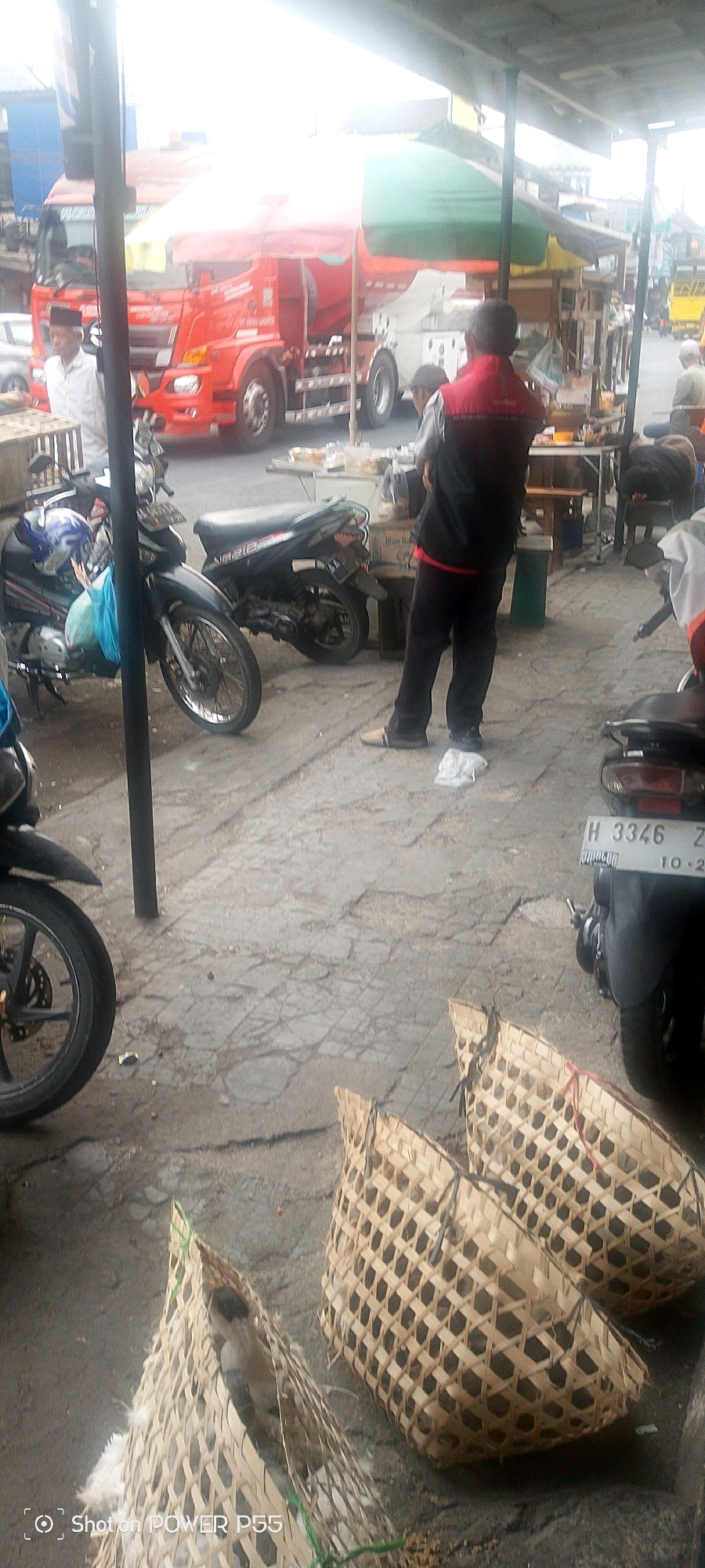 The image depicts an outdoor market scene, likely in a city. In the foreground, there are two woven bamboo baskets, one of which contains a bird. Several motorcycles are parked around the scene. In the background, a large red truck is visible, along with market stalls and awnings. People are present, including a man wearing a red vest and dark pants standing near a stall, and another man in a white shirt near the truck. The ground appears to be paved with rough concrete.