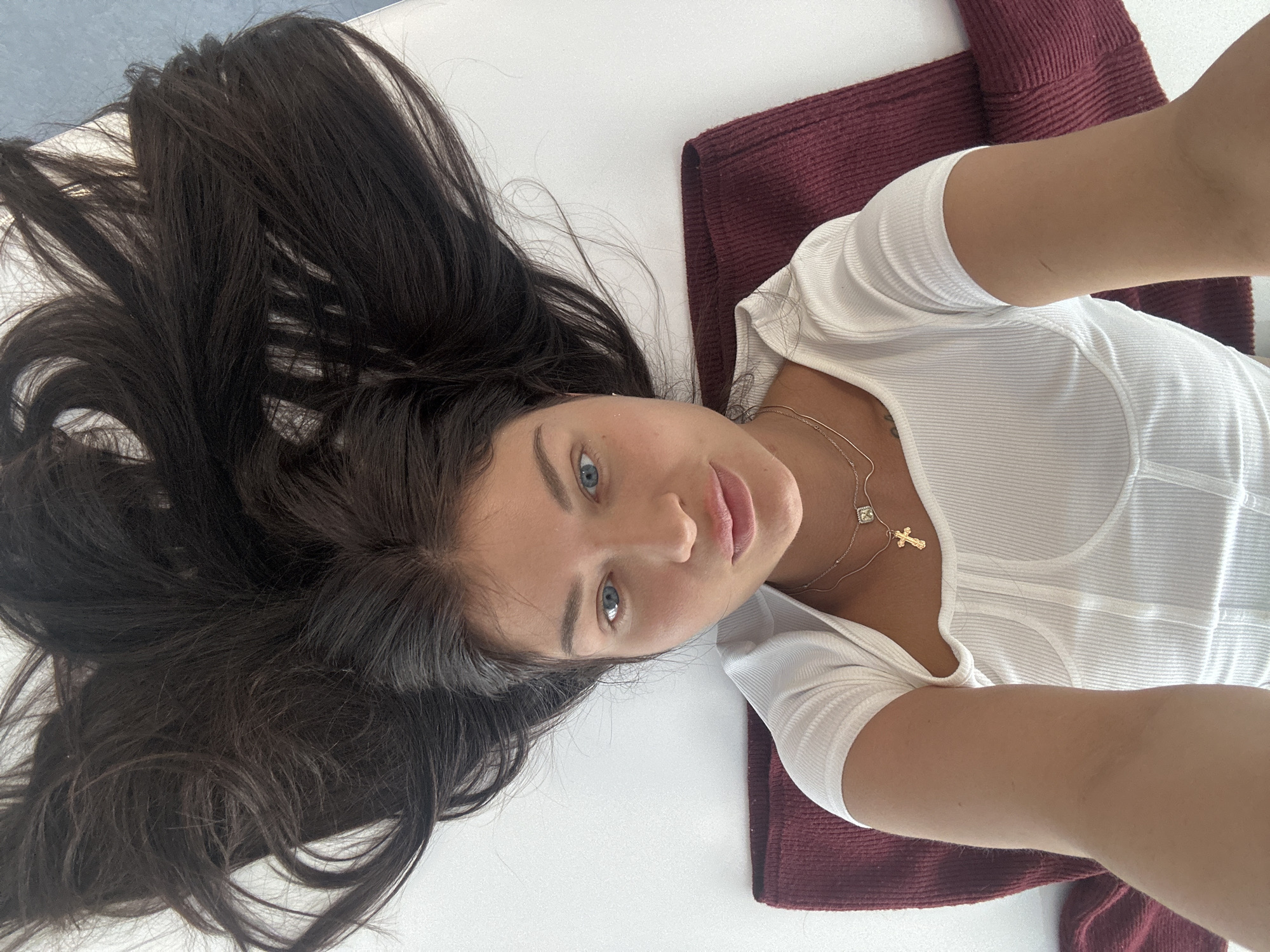 A young woman with long, dark, wavy hair and striking blue eyes is depicted in a close-up, top-down view, likely a selfie. She is lying on her back, looking directly at the camera with a calm, neutral expression. Her dark hair is fanned out around her head against a white surface, possibly a bed or floor.
She is wearing a white, ribbed, short-sleeved top. Around her neck, she has two delicate gold necklaces: one features a small, square, clear stone pendant, and the other a small gold cross pendant. Her skin appears smooth and lightly tanned.
To her right, a segment of dark reddish-brown, textured fabric is visible, possibly a blanket, mat, or garment, adding a subtle color contrast to the predominantly white background. The lighting is soft and even, suggesting an indoor setting without harsh shadows, potentially during the daytime. No specific location cues are discernible within the image itself to confirm the given context of Khimki, Russia, and no text is visible. The image captures a serene and candid moment.