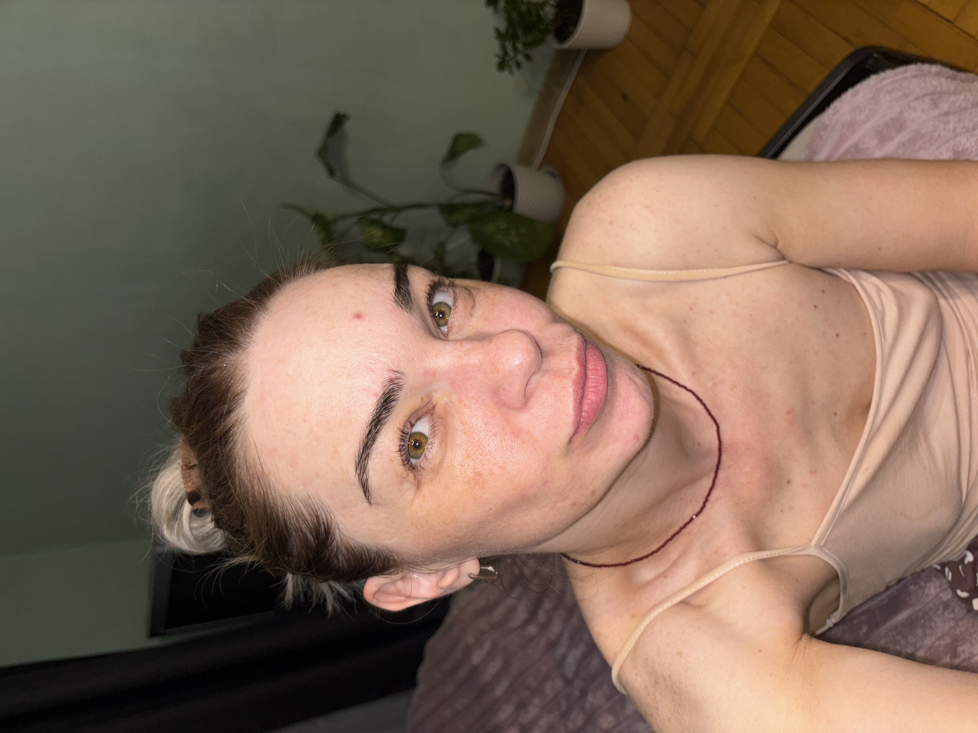 A young woman with blonde hair pulled up in a messy bun is looking at the camera. She is wearing a beige tank top and a red beaded necklace. The background shows a wall with some plants and wooden floors. The overall lighting suggests an indoor environment, possibly a bedroom. There are no explicit indications of the city or country, and therefore no specific location in "Unknown, Russia" can be determined.