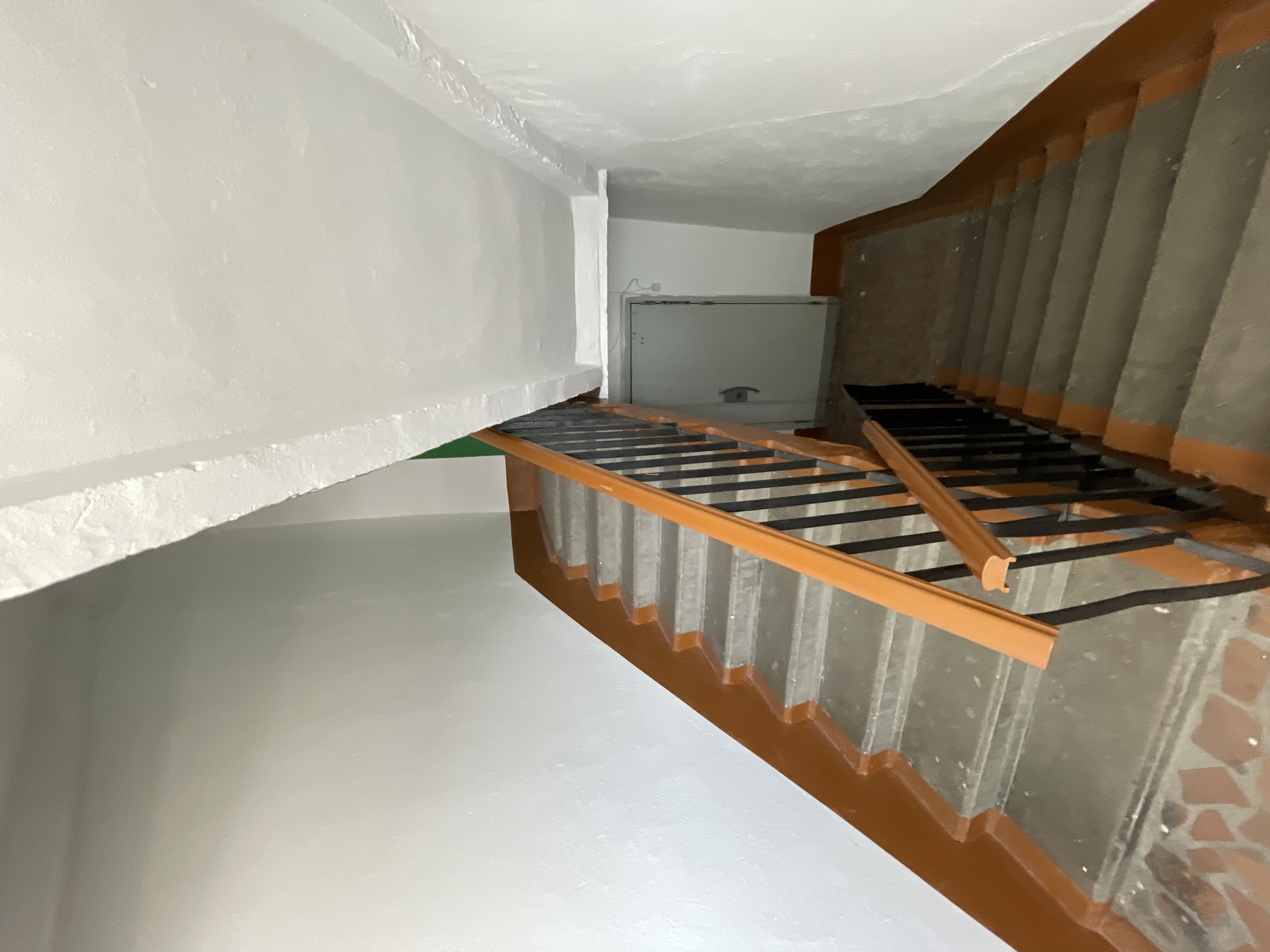 The image shows a concrete staircase in what appears to be an apartment building's entryway in Spassk-Dal'nij, Russia. The staircase has a brown painted trim along the edge of the steps and a black metal railing with a wooden handrail. There is a metal door visible at the bottom of the stairs. The walls are painted white. The lighting suggests it might be daytime, but the interior is dimly lit.