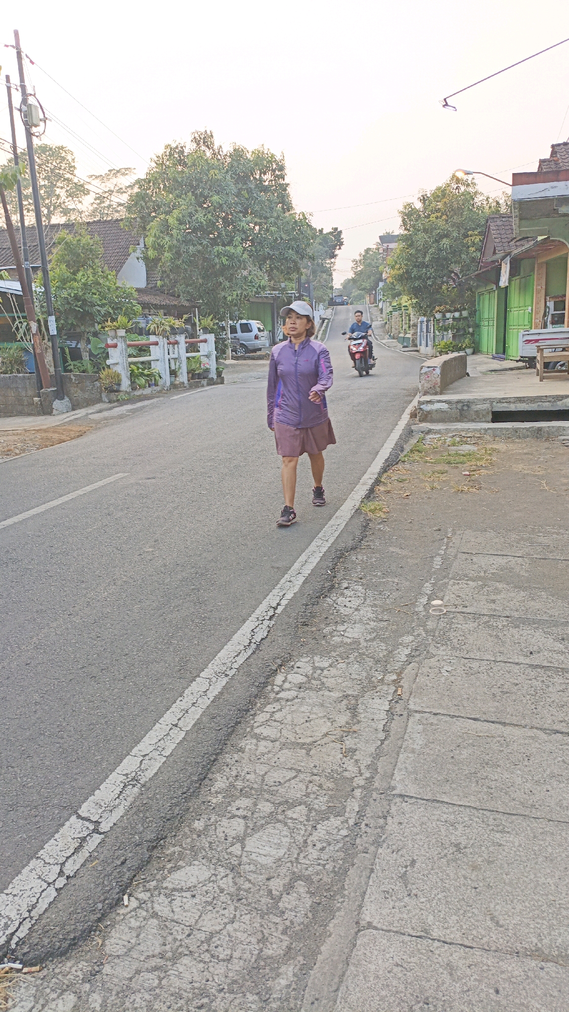 A woman in a purple jacket and skirt walks on the right side of a two-lane asphalt road. She is wearing a gray cap and dark shoes. A man on a red motorcycle rides in the same direction, further down the road. The road is lined with houses and trees. The sky is overcast. The scene appears to be in a residential area. This photo was taken on Jalan Argowiyoto, Salatiga, Indonesia.