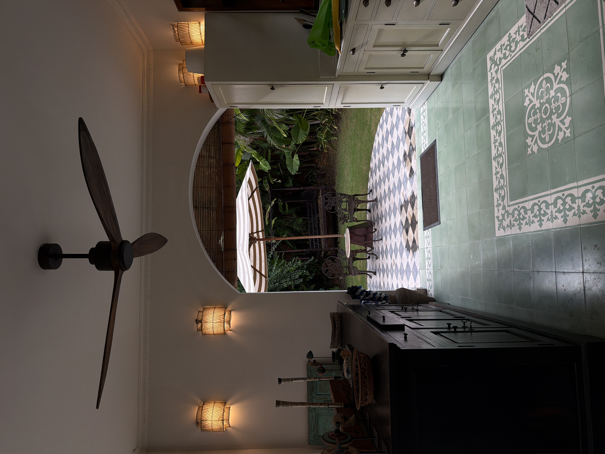 The image shows a well-lit interior space with an archway leading to an outdoor patio and garden. The interior features a dark wooden cabinet on the left and a white cabinet on the right, both with multiple drawers and doors. Above, a dark wooden ceiling fan with three blades is visible. Two rattan wall sconces with illuminated bulbs are mounted on the left wall. The floor in the interior is tiled with large, pale green squares.
Through the archway, the patio is tiled with a checkerboard pattern of black and white squares. On the patio, there is a small round table with two ornate chairs. An umbrella with white and beige stripes stands open near a wooden swing set in the background. The garden beyond is lush with tropical foliage. The scene suggests a residential setting in North Kuta, Indonesia, characterized by its tropical climate and architectural style. There are no people or animals visible in the image.