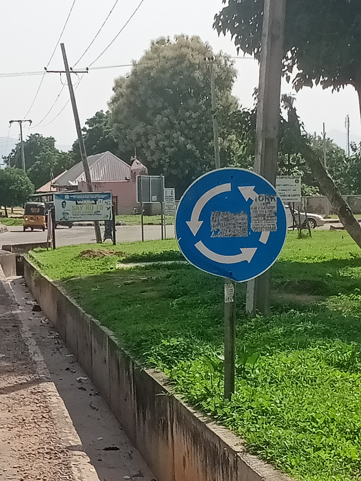 The image depicts a roundabout traffic sign in Jalingo, Nigeria. The sign, circular and blue with white arrows, indicates a mandatory direction of travel around the roundabout. A wooden utility pole stands behind the sign, with power lines visible against a bright sky. To the left of the sign, a yellow auto-rickshaw is seen on the road, and a billboard with images of two men and text including "WABA SAY NO TO..." is also visible. The area is lined with a concrete barrier and overgrown grass. In the background, buildings and trees can be seen, with a glimpse of mountains on the horizon. The scene appears to be a street corner or intersection within the city.