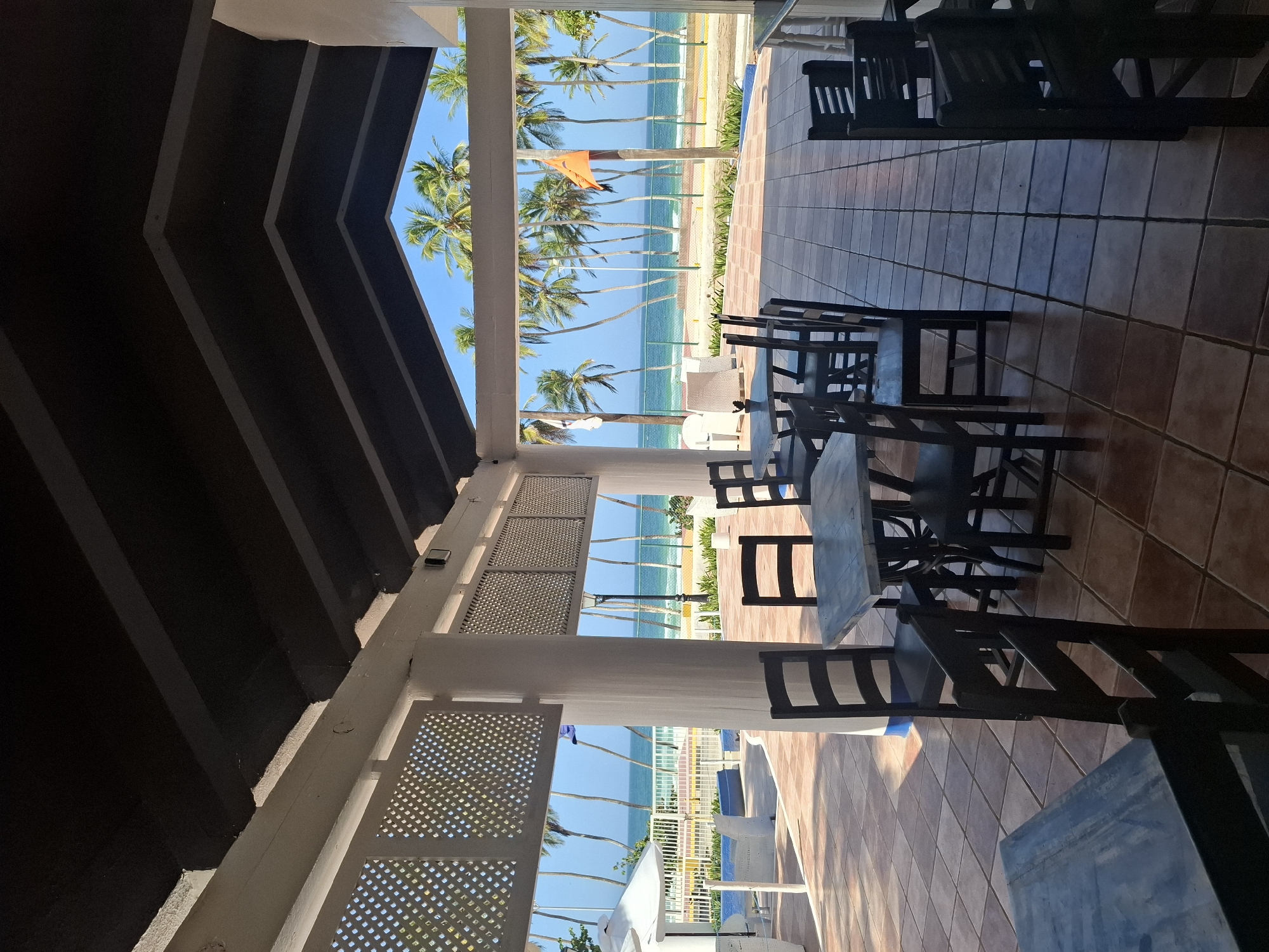 The image captures a tranquil scene from an outdoor dining or seating area, looking out towards a tropical beach. The perspective is from within a shaded structure, likely a pavilion or open-air restaurant.
In the foreground and midground, numerous dark wooden tables and chairs are arranged on a light brown tiled floor, with distinct shadows cast by the furniture and the overhead structure, suggesting bright sunlight. The structure itself features dark, angled ceiling beams and white support pillars. Decorative white lattice panels frame the sides of the viewing area, adding to the architectural detail.
Beyond the shaded area, a vibrant tropical landscape unfolds under a clear blue sky. A prominent line of tall palm trees stretches across the frame, separating the seating area from the sandy beach. An orange flag on a pole stands out against the green foliage and blue sky, located on the beach.
Further in the distance, the serene turquoise waters of the ocean meet the horizon. Hints of white and blue lounge chairs and other resort structures can be seen along the beachfront. The strong natural light and long shadows indicate a sunny day, likely late morning or early afternoon. No people are visible in the scene, contributing to a sense of peaceful quietness. No visible text is present. The setting is consistent with a resort location like Playa El Agua, Venezuela, as provided by the context.