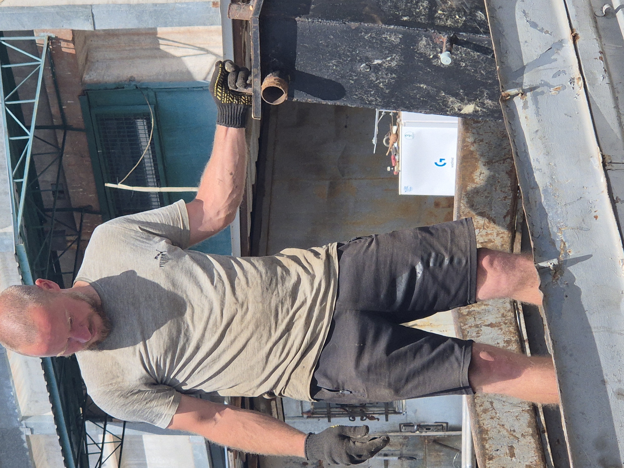 A bald, fair-skinned man with a beard and a light-colored t-shirt and dark shorts stands in a weathered metal container. He wears black work gloves with yellow accents on his right hand, which is resting on the edge of another metal structure. His legs are visible up to his knees, and he appears to be inside the container. Behind him, a white refrigerator with a blue logo is visible, along with other assorted items and structures that suggest an industrial or outdoor work environment. The scene is lit by sunlight, casting shadows. The man is in Pidhaitsi, Ukraine.