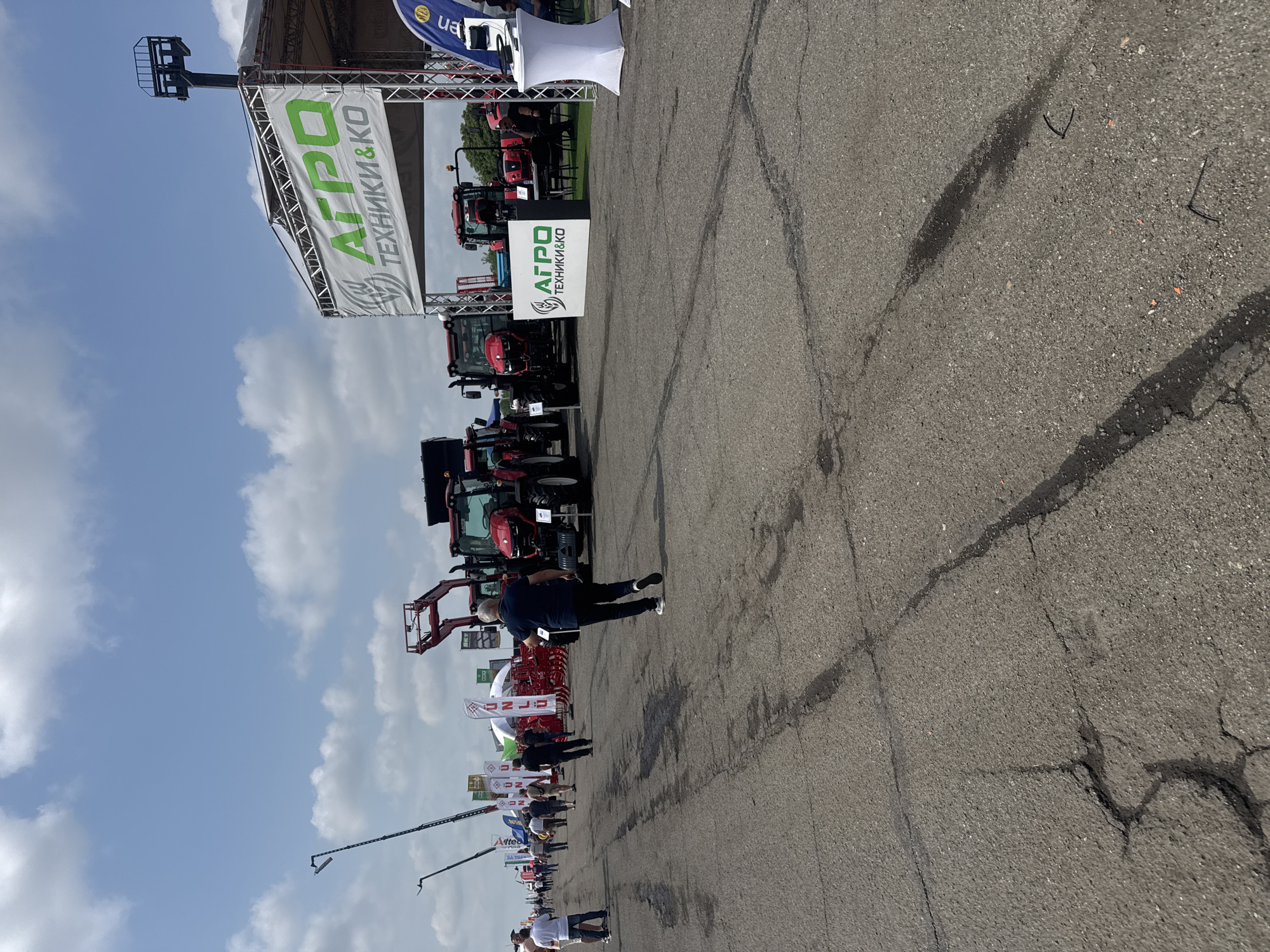 The image, when viewed with correct orientation (rotated 90 degrees clockwise), depicts an outdoor agricultural machinery exhibition or trade fair in Stara Zagora, Bulgaria. The event is set on a vast, cracked asphalt surface under a bright blue sky with scattered white clouds, indicating daytime.
Numerous red and black agricultural tractors and other farm implements are prominently displayed across the paved area. These machines vary in type, including several tractors, one fitted with a front-end loader attachment, and other machinery like sprayers or cultivators.
Many people are visible, casually dressed, walking among the exhibits and observing the machinery. One man in a dark blue shirt and black pants is distinct in the midground, seen from behind, facing the displays. Other attendees are scattered throughout the scene, engaging with the exhibition.
A large green and white banner suspended from a metal truss structure prominently displays the text "АГРО ТЕХНИКИ&КО" (AGRO TECHNIKI&KO) along with a stylized wheat stalk logo. A smaller, freestanding sign with the identical "АГРО ТЕХНИКИ&КО" branding is also visible near the machinery. Other partially visible banners include "ua" on a blue background and "BKT", suggesting additional exhibitors or sponsors.
The ground features visible cracks and some dark, wet-looking stains on the asphalt. In the background, there's a white canopy or tent structure, typical of an outdoor exhibition. The scene conveys an active and well-attended industry event focusing on agricultural technology.