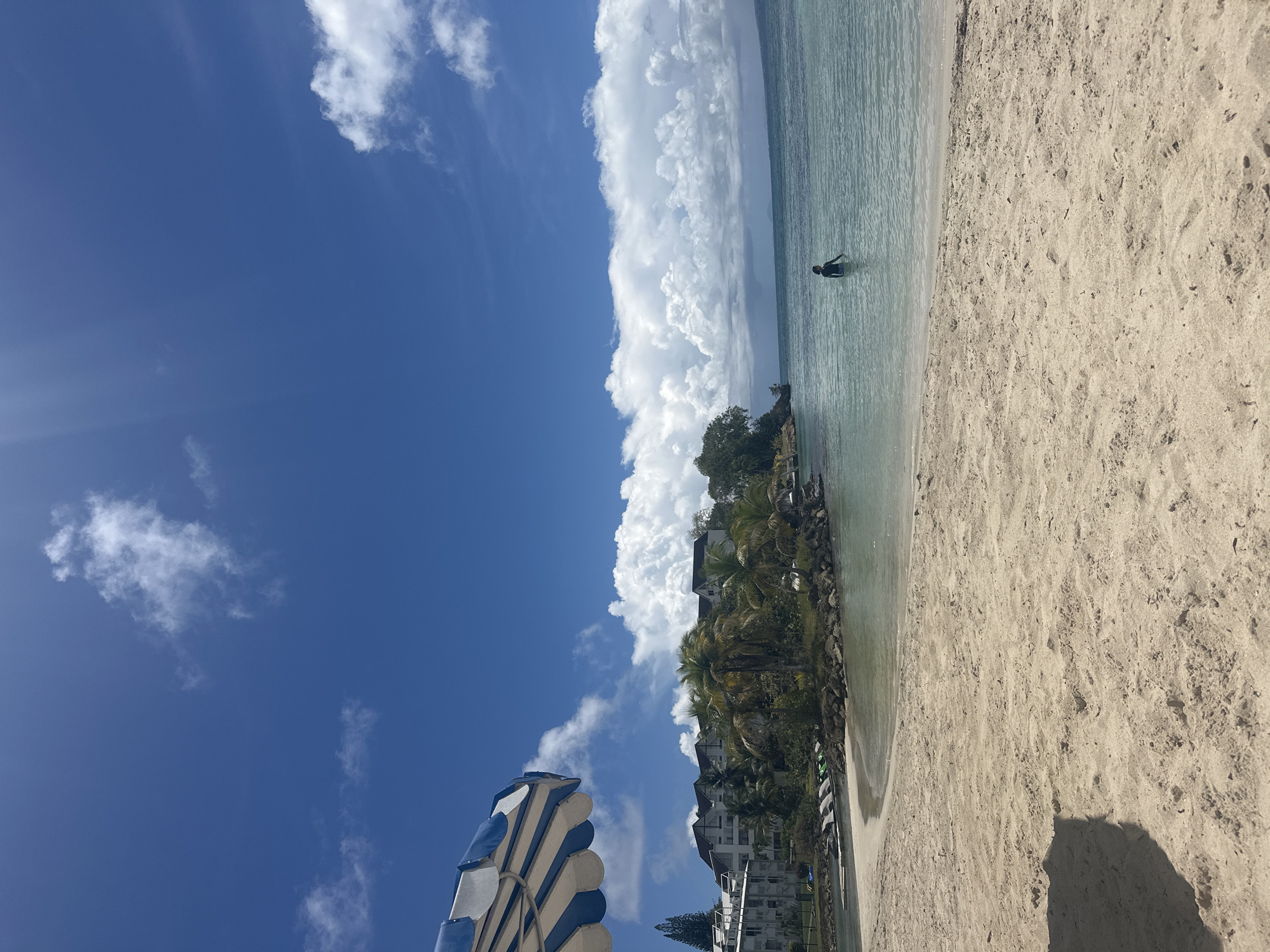 The image depicts a beach scene in Le Gosier, France. A person is standing in the water, near a rocky outcropping with vegetation. The beach is sandy, and there is a blue and white beach umbrella in the foreground. The sky is blue with large, white cumulus clouds. Buildings are visible behind the vegetation.