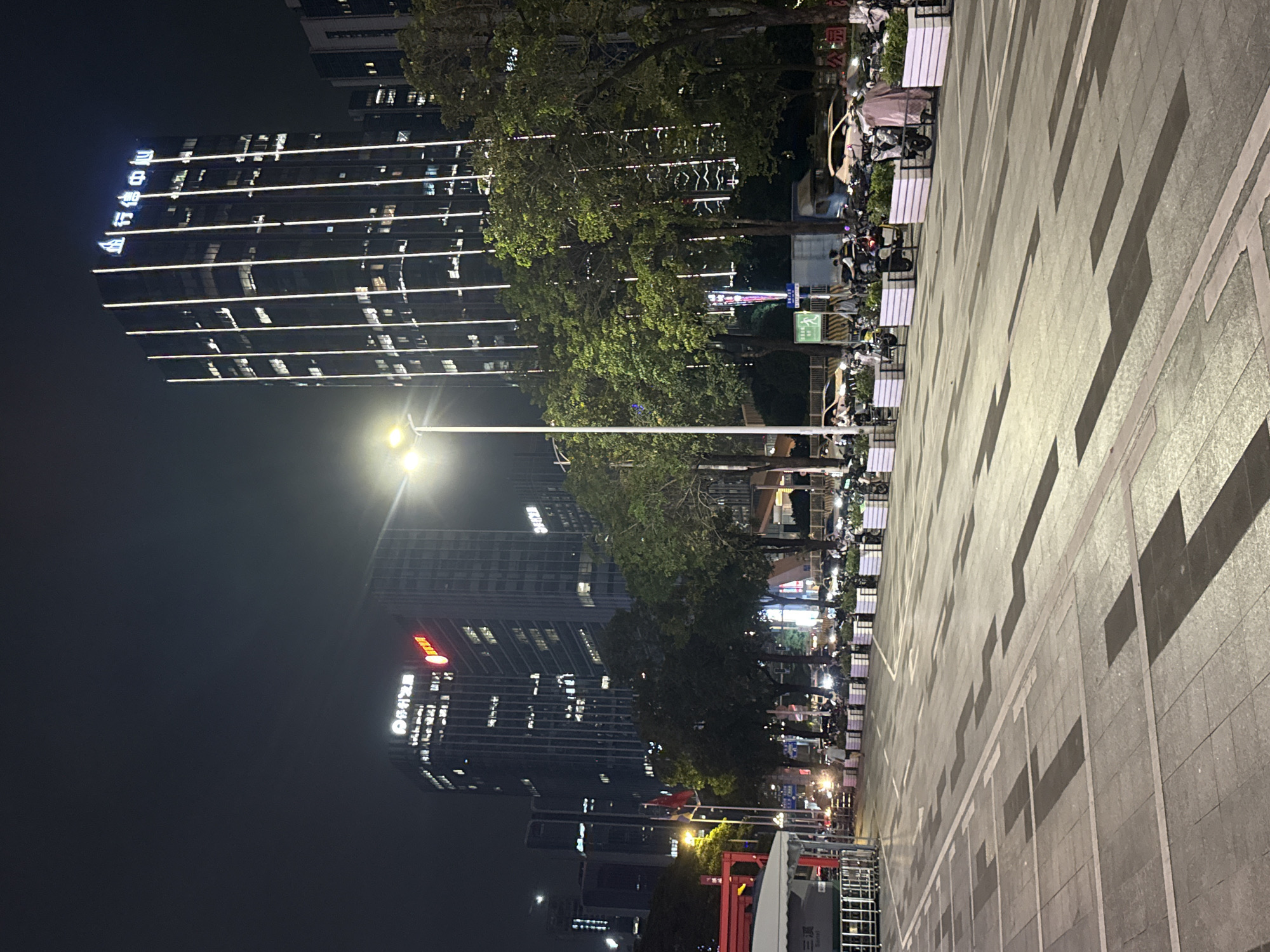 The image depicts a street scene at night in Tianhe District, China. A tall, modern skyscraper, illuminated by vertical white lights and topped with blue signage, dominates the background. To the left, another skyscraper is visible with prominent red and white signage. The street is paved with large, light-colored tiles, featuring a geometric pattern of darker rectangles and white lines. Along the side of the street, several rows of white planters filled with greenery separate the pedestrian walkway from the road. A variety of parked motorcycles and scooters are visible in front of the planters. A few cars are present on the road, with their headlights on. A street light with a bright bulb illuminates the foreground. In the distance, trees line the street, and the lights of other buildings are visible. A person in a light-colored garment is walking away from the camera, towards the right side of the frame, near the planters. A small red and white structure with a canopy is visible on the far left, possibly a kiosk or a temporary barrier. The overall atmosphere is that of an urban environment at night, with clear visibility due to artificial lighting.