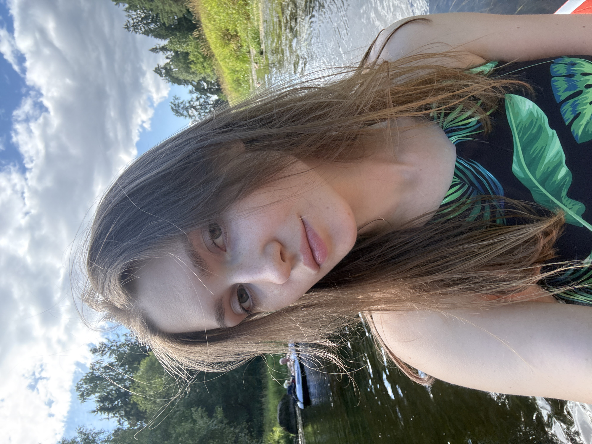 In a selfie taken from a low angle, a young woman with long, light brown hair is prominently featured, her face filling the foreground. She is outdoors, on a body of water, with a backdrop of trees and a cloudy sky. She is wearing a black swimsuit with a green palm leaf pattern. Her gaze is directed towards the camera. In the background, further down the river, two individuals are visible in an inflatable raft. The water is calm, with reflections of the sky and surrounding foliage. The overall impression is one of a leisurely outdoor activity on a summer day. There is no identifiable signage or architecture within the frame to indicate a specific location within Старосиверская, Russia.