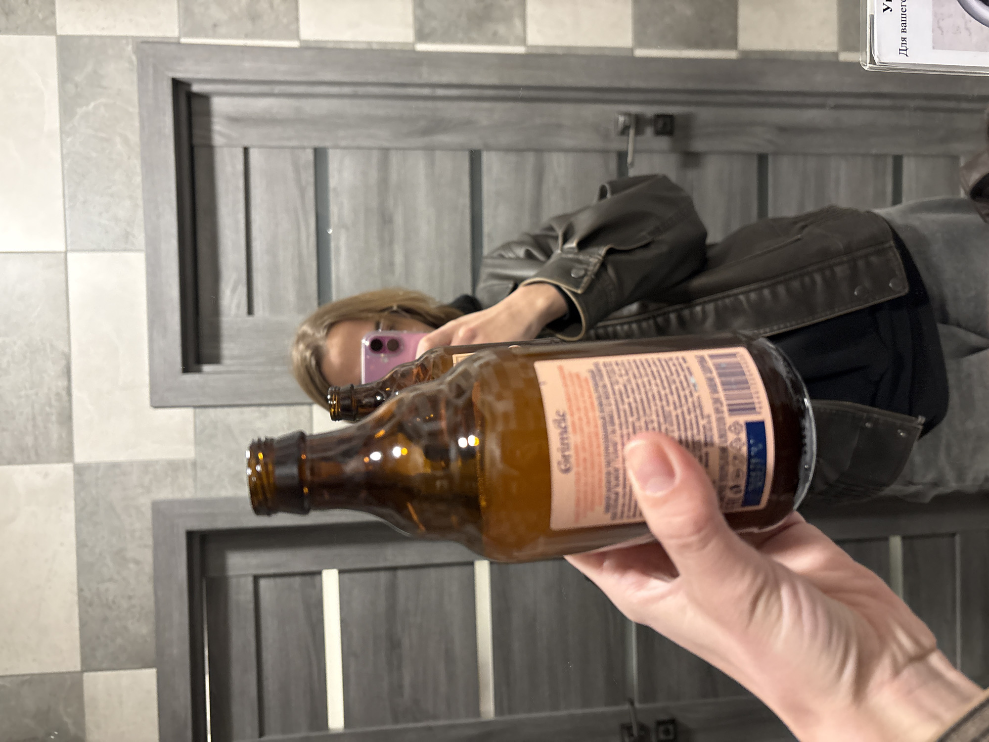 A person in a brown leather jacket is holding a brown glass bottle labeled "Grindle" in front of a mirror. The person is reflected in the mirror, taking a photo with a pink phone. The background includes a tiled wall and a gray wooden door with vertical grooves. The scene is presumably inside a bathroom based on the tiled wall and mirror. There is no explicit information to conclude the location as Ust-Ilimsk, Russia.