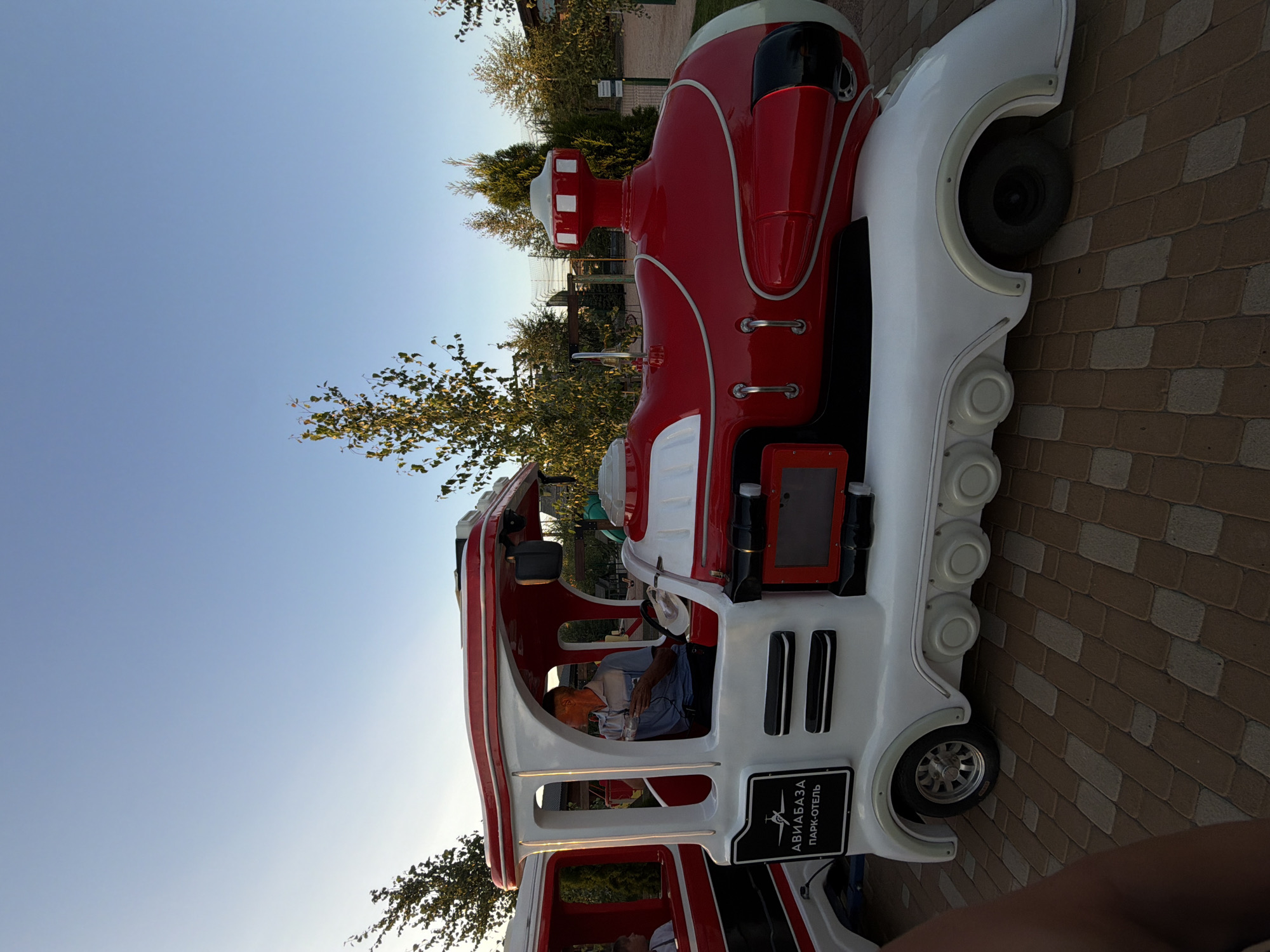 The image captures a red and white miniature train with a driver. The train is on a paved walkway in an outdoor setting. The driver, a middle-aged man wearing a light blue collared shirt and dark pants, is seated in the front car, holding a clear plastic container and looking to his left. A white hat rests on the steering wheel. The train has a prominent red boiler-like section and white detailing. On the side of the front car, a black rectangle contains a logo with a plane and text in Russian that reads "АВИАБАЗА ПАРК-ОТЕЛЬ" (Aviabaza Park-Hotel). Behind the train, there are trees and a clear blue sky. The ground is paved with interlocking bricks in a pattern of tan and off-white. The image appears to have been taken during daylight hours. The location is identified as the Aviabaza Park-Hotel in Kamyshin, Russia.