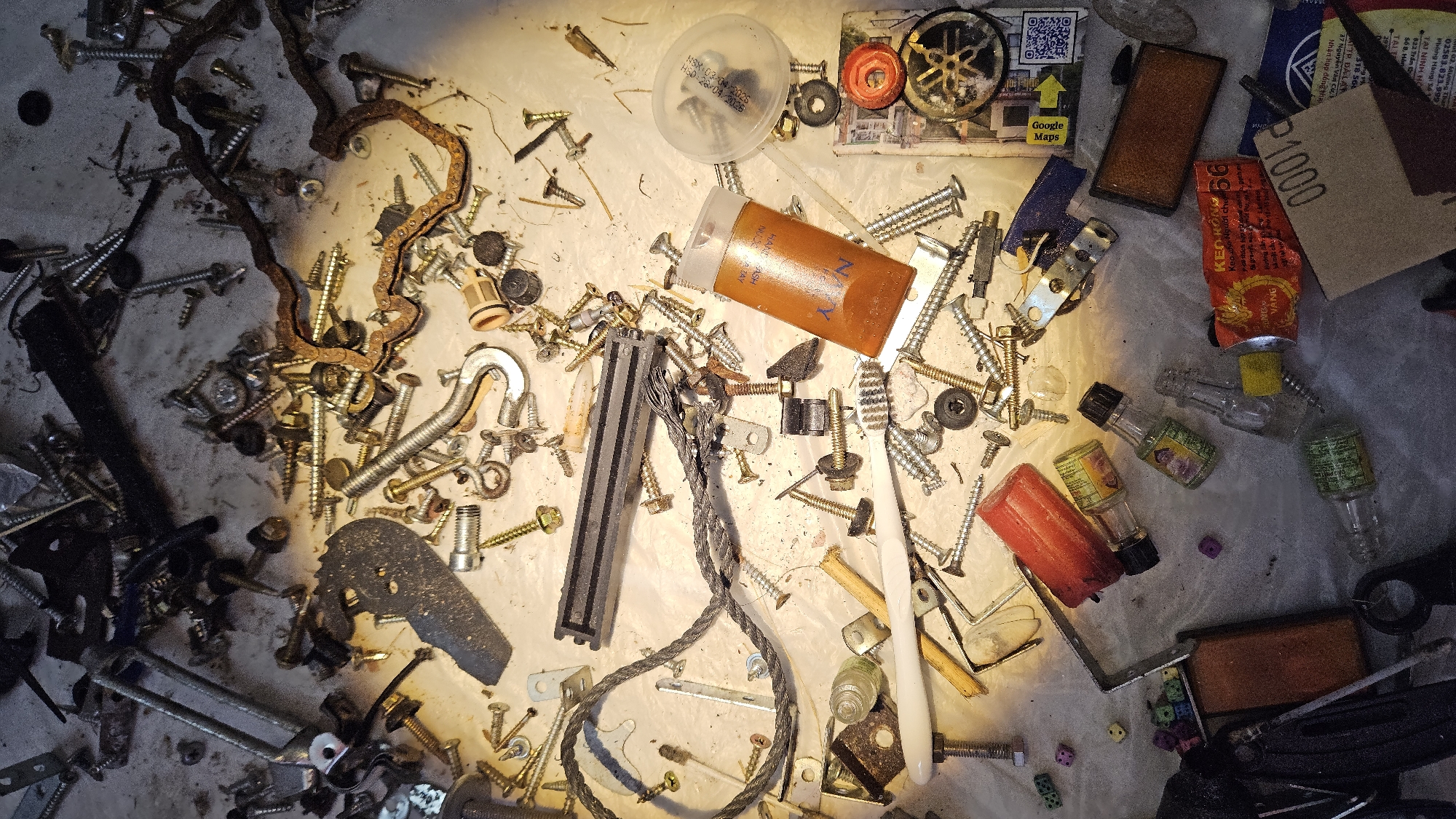 The image presents a top-down view of a cluttered, light-colored surface, possibly a workbench or floor, illuminated by a direct, warm light source that creates distinct shadows. The scene is filled with a miscellaneous collection of small objects, indicating a setting like a workshop or storage area, likely in Cam Đức, Vietnam.
The primary subjects are numerous metal fasteners, including a wide variety of screws (phillips, slotted, hex head), nuts, bolts, and washers, many showing signs of wear or rust. Other hardware items include metal brackets, a spring, a U-shaped hook, a braided metal cable, and a rusty, wavy chain-like component or gasket.
Among the clutter are several containers and personal items. A clear plastic container with a lid displays "NSX 03/04/2025" and "HSD: 28/04/2026". An orange cylindrical bottle is partially labeled "HAND WASH" and "NYV". A red tube, likely an adhesive, shows "KEO 56", "Keo", and "VANG". Several miniature glass bottles with green labels and black caps are scattered across the surface. Other items include a white toothbrush, multiple small, colorful dice (purple, green, orange), and a piece of sandpaper marked "P1000".
Visible text on a piece of paper includes a "Google Maps" logo, a QR code, and extensive Vietnamese text: "29", "02583.823.999", "Phòng Hàng Số", "TẠI NINH HO", "822 Trần", "Số 8, Hồng", "2. Mương Than", "318 564", "37 Nguyễn Văn Cư", and "Nhận hợp đồng thai". A stylized Yamaha logo is also partially visible on another item. No specific activity or event is taking place; the scene suggests a collection of assorted items, possibly awaiting organization or disposal.