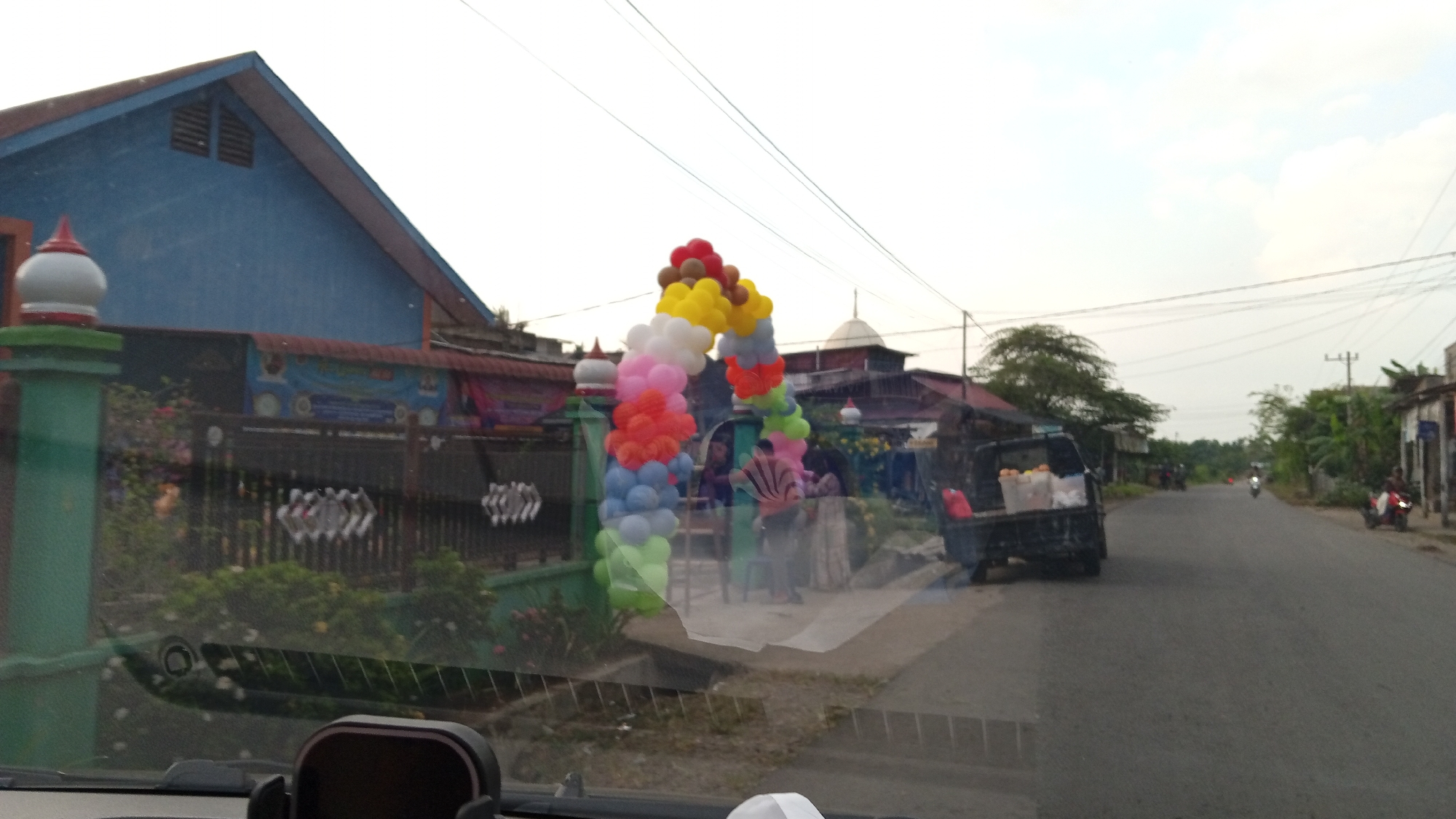 In Kilometer Delapan, Indonesia, the image captures a street scene with a blue building on the left, featuring a pitched roof and vent grilles. In front of the building, a green gatepost with a white and red finial stands, partially obscured by a reflective surface, possibly a car windshield. A balloon arch, adorned with multiple colorful balloons in red, yellow, white, pink, orange, blue, and green, spans across the road, framing the entrance.
Behind the balloon arch, a dark fence with a decorative pattern encloses a green area with foliage and flowers. A banner is visible on the fence. In the background, several buildings with pitched roofs are present, along with a mosque-like dome.
On the right side of the street, a dark-colored pickup truck is parked, its bed filled with items, including what appear to be boxes and a red bag. Further down the road, several motorcycles are visible, with one person riding a red motorcycle on the right side and another on a white motorcycle in the distance. The road is paved, and overhead power lines are present. Trees and vegetation line the sides of the road, and the sky is overcast with clouds. A dark object, possibly a car mount, is in the foreground at the bottom center of the image.