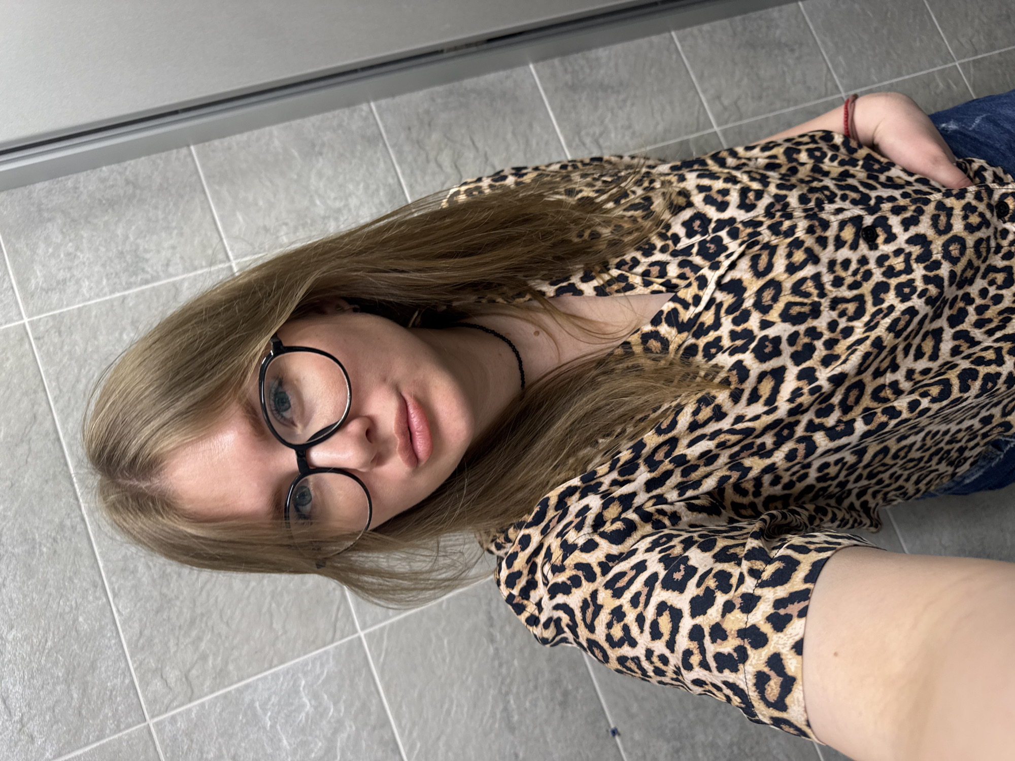 A woman is indoors, likely in Perm, Russia, based on context. She is wearing glasses and a leopard print shirt. She is also wearing a dark necklace and a red bracelet. She is likely wearing denim jeans. The floor is tiled and gray.