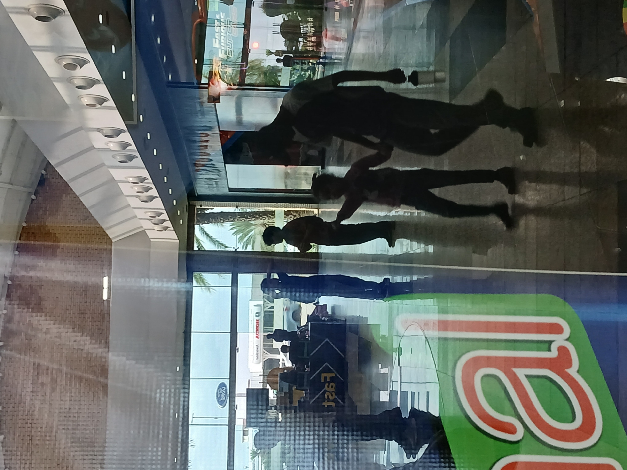 The image captures a scene inside the Ciudad Alta Vista II Shopping Mall in Ciudad Guayana, Venezuela, as seen reflected in a glass surface. Three indistinct human figures are present, appearing as reflections. In the background, through the glass, a store or establishment is visible with an "ACTIROAR" sign and other business signage. A "Ford" logo is also present. The foreground is dominated by a distorted partial view of what appears to be a large logo with green and red colors. The lighting is artificial, with a row of recessed lighting fixtures visible in the ceiling. The overall scene suggests an indoor environment during daylight hours.