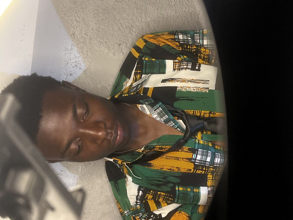 The image depicts a young adult Black male, centrally positioned in an interior setting in Bar Gada, Nigeria. He is shown from the chest up, with his head slightly bowed and eyes directed downwards. He wears a long-sleeved, collared button-down shirt featuring an abstract pattern of vertical and horizontal brushstrokes and grid motifs in shades of green, yellow, black, and white. A thin, silver-colored ball chain necklace is visible around his neck, beneath an untied black tie draped loosely around his collar. The background consists of a light-colored, textured wall, possibly stucco. An out-of-focus, dark gray object, appearing metallic or plastic, occupies the upper left portion of the frame, possibly a light fixture or a photographic accessory. The lower section of the image is obscured by a dark, out-of-focus foreground element. The subject is still, with no apparent action or interaction with others.