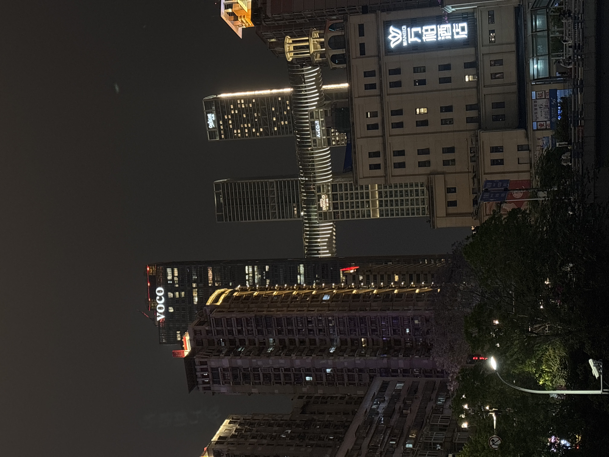 This media file captures a nighttime cityscape, likely in Yuzhong District, China, characterized by numerous illuminated tall buildings and skyscrapers under a dark, clear sky. The image is taken from a street level perspective, looking up at the urban environment.
Several prominent buildings feature bright signage. On the left, a tall building displays "VOCO" vertically in white letters, indicating a hotel. In the center, a striking building with horizontal light bands features "ASCOTT" and "INDIGO" signs, suggesting serviced residences or hotels. Another modern building nearby has "Calltrust" visible. Towards the right, a multi-story building features the prominent "WANBO" and Chinese characters "万宝" (Wànbǎo), possibly a department store or commercial complex. Below this, smaller signs with Chinese characters like "今乐游园" (Jīnlè Yóuyuán) and "旅" (lǚ, meaning travel) are visible, indicating local businesses or advertisements.
The buildings exhibit various architectural styles, from modern glass facades to more traditional structures with numerous windows, many of which are lit, creating a scattered pattern of light. In the foreground on the right, dark green trees and foliage partially obscure some lower buildings. A street light with a bright white lamp illuminates a section of the foreground, and a red traffic light signal is visible above it. A partially obscured "no parking" sign can be seen near the bottom right. The overall impression is one of a bustling, active urban area after dark.