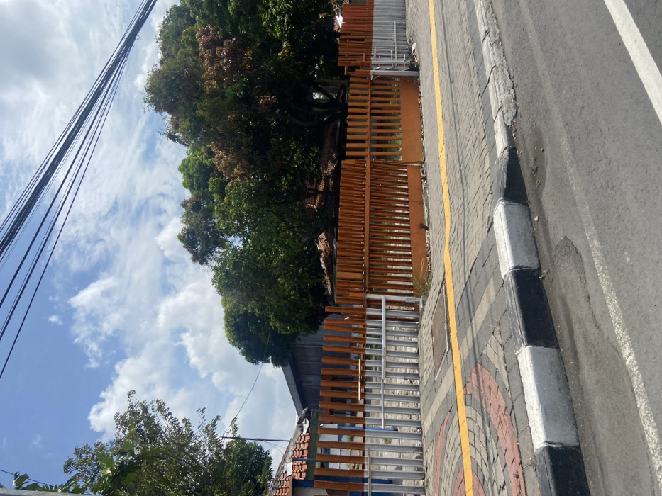 The image shows a street in Mojokerto, Indonesia (latitude: -7.481, longitude: 112.44). The street is paved with asphalt and has a sidewalk with a yellow painted edge. The sidewalk is made of concrete blocks and a curb. There is a wooden fence with orange slats on the right side of the image. The fence is partially open. In the background, there is a group of trees and a blue sky with white clouds. There are multiple electrical wires running across the sky.